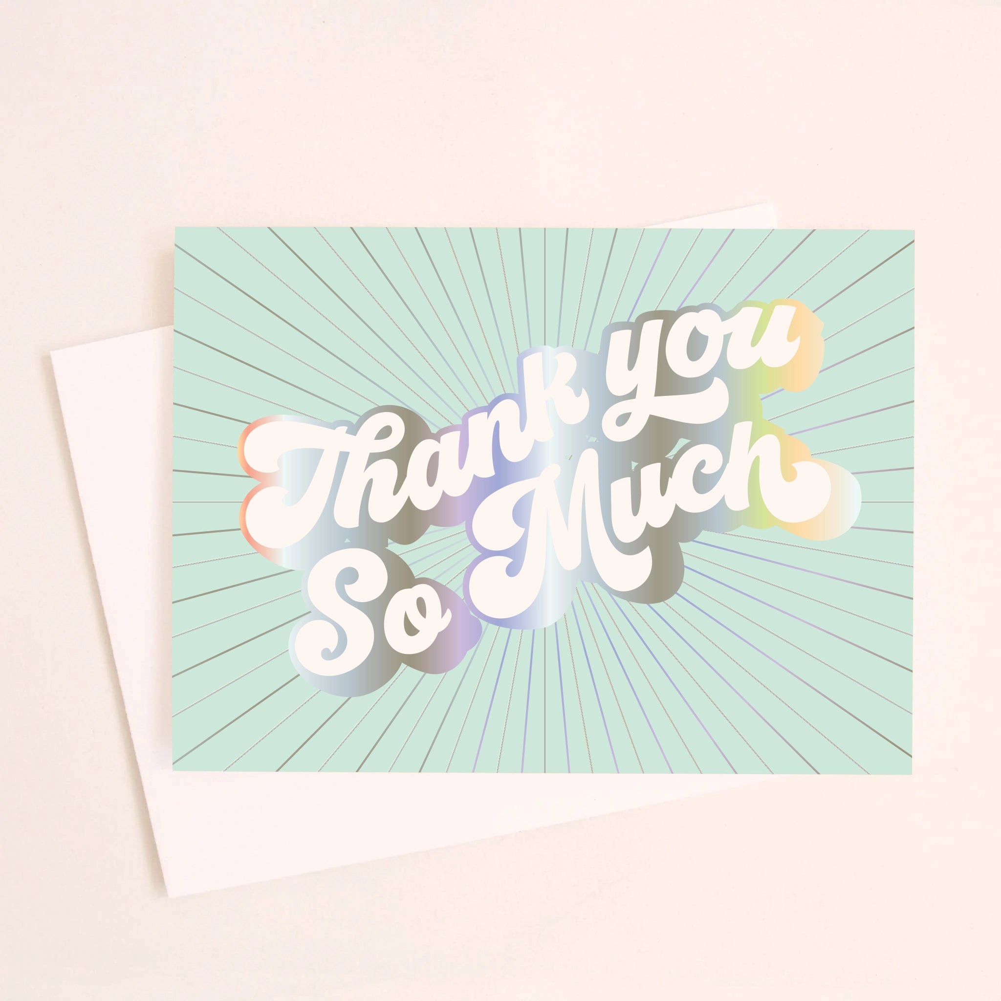 On an ivory background is a light teal greeting card with white, holographic outlined text that reads, &quot;Thank You So Much&quot;.