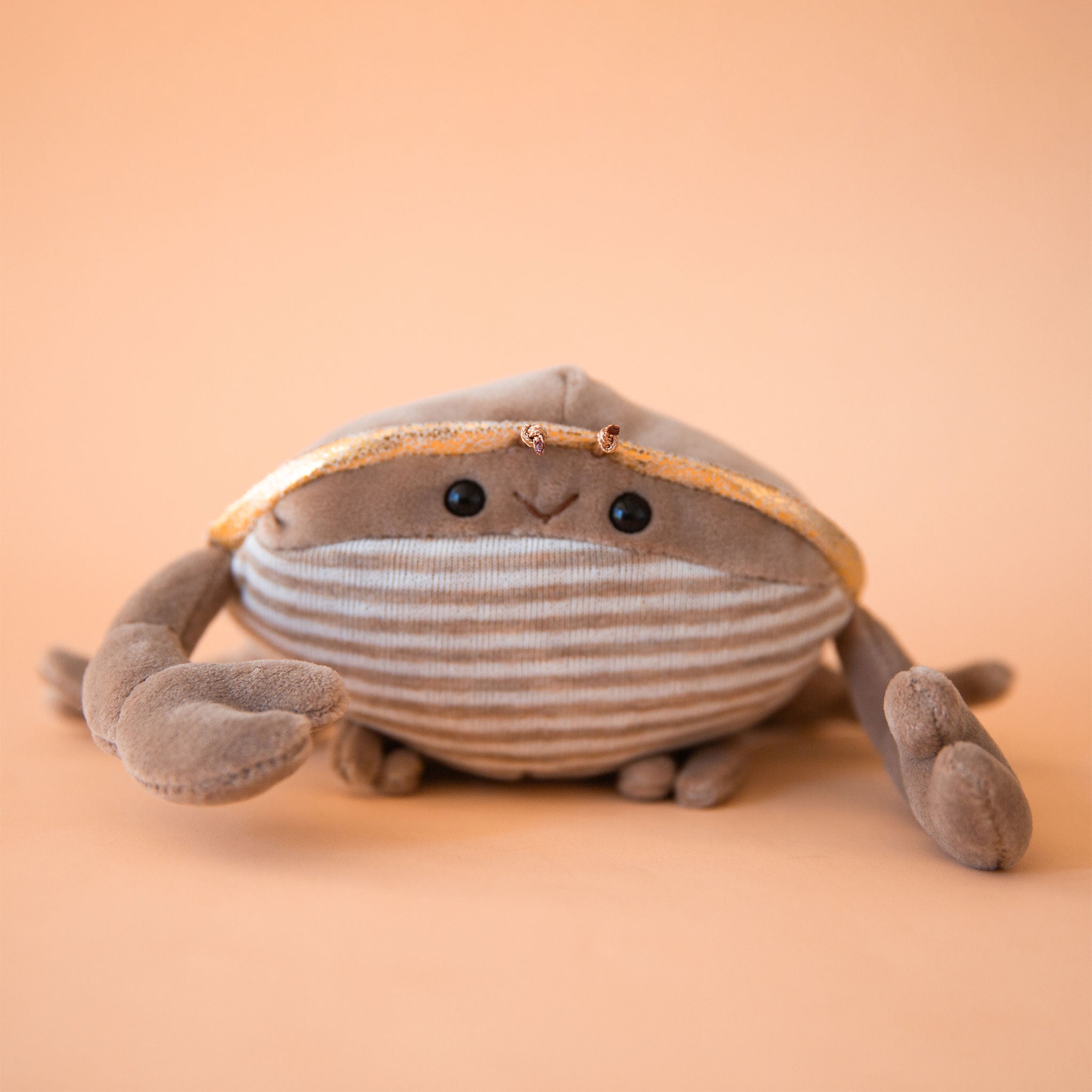 On a tan background is a grey ish brown crab stuffed toy with a sweet smiling face.