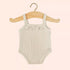 A beige colored doll onesie outfit hangs on a wooden hanger.  The outfit is a light beige color with spaghetti straps and a ruffle at the neck.