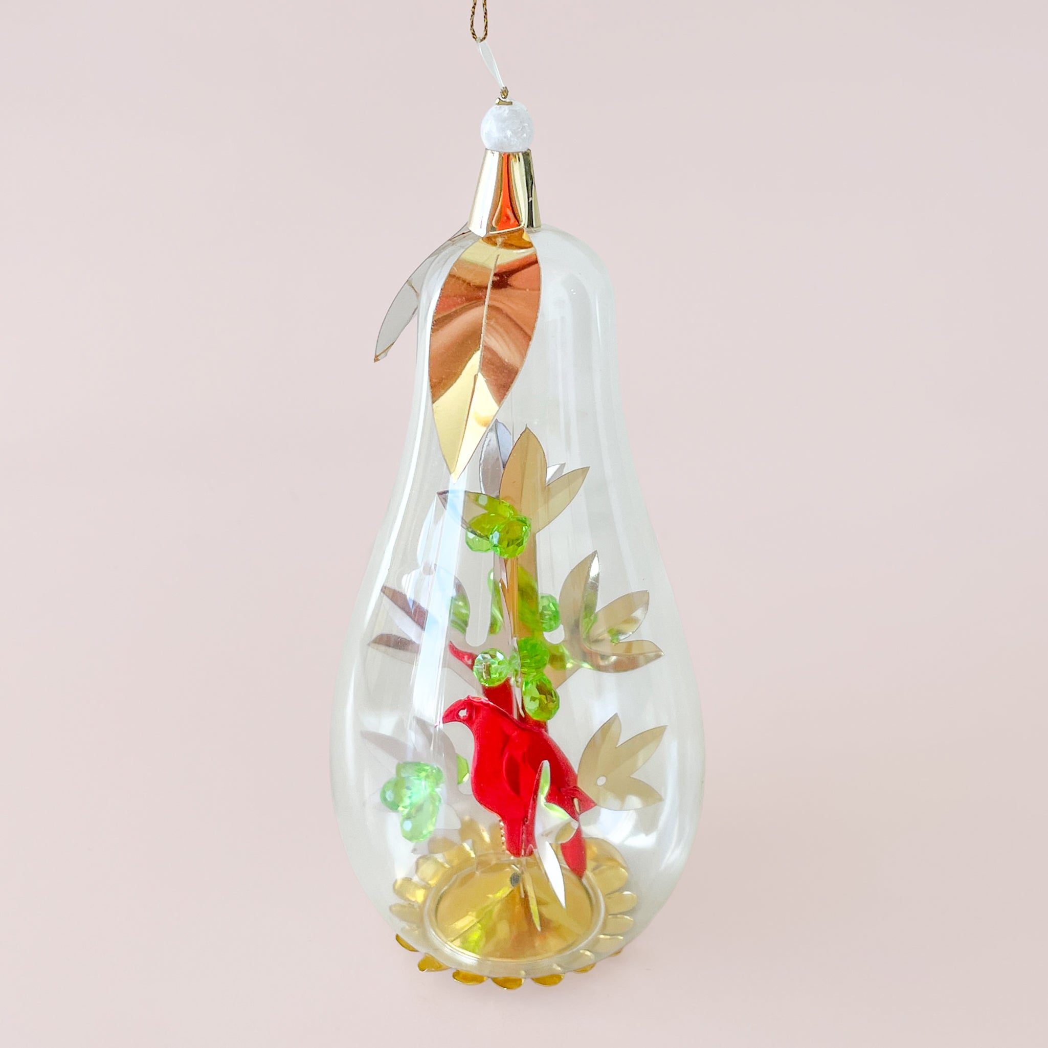 A clear, pear-shaped glass ornament hangs against a pale pink background, decorated inside with a small red bird, green berry clusters, and gold leaf accents.