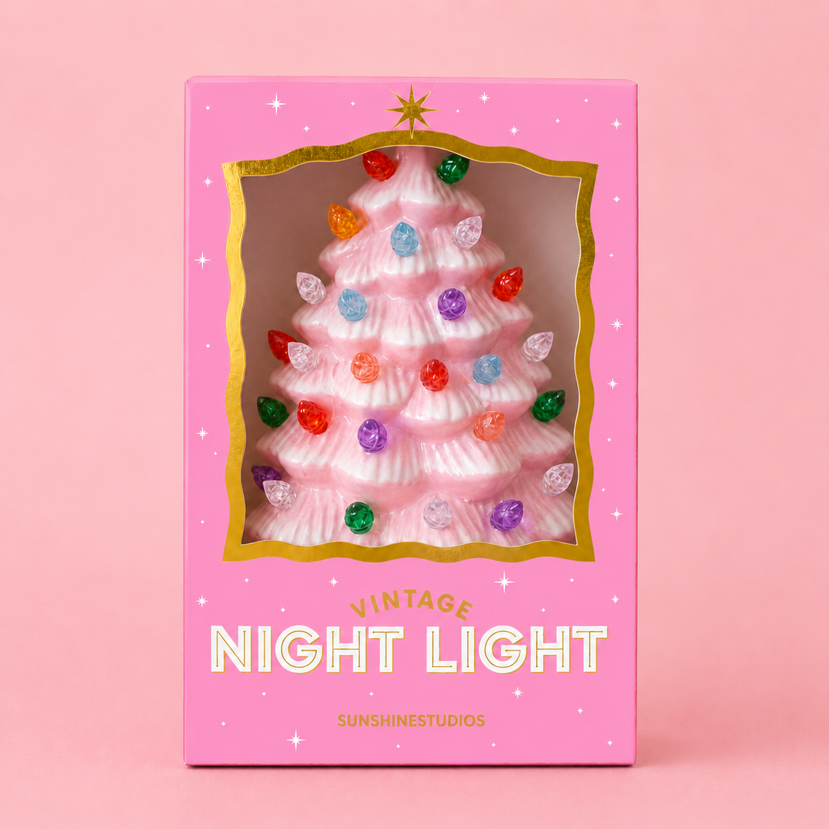 A pink Christmas tree-shaped night light in its original packaging, featuring a clear window that shows the light on, with colorful bulb accents. The box is pink with gold trim and white text reading 'VINTAGE NIGHT LIGHT' and 'SUNSHINESTUDIOS'.
