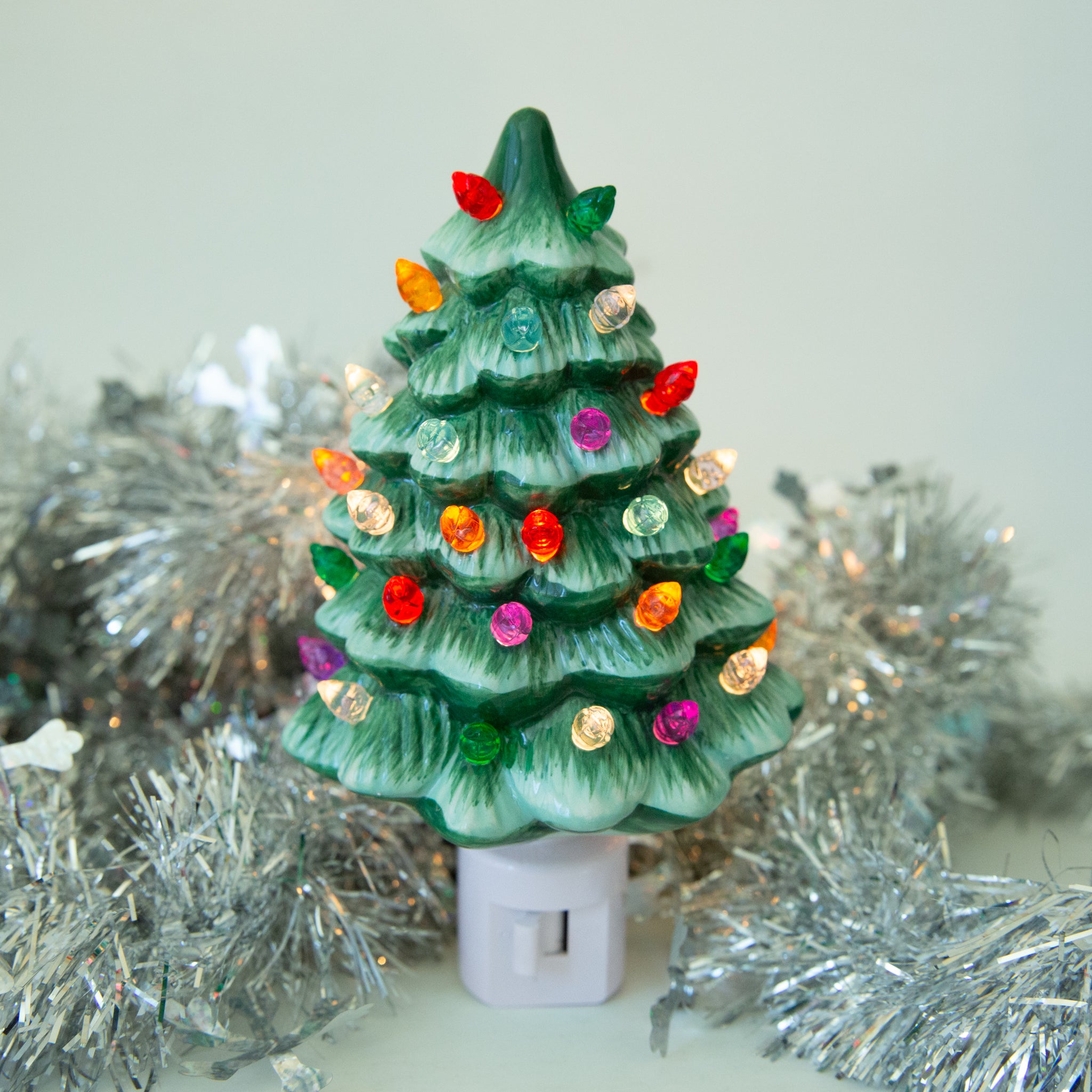 A ceramic Christmas Tree shaped night light sits in middle of the page with bright colorful lights.  It has a white plug at the bottom.  The tree is a green color.