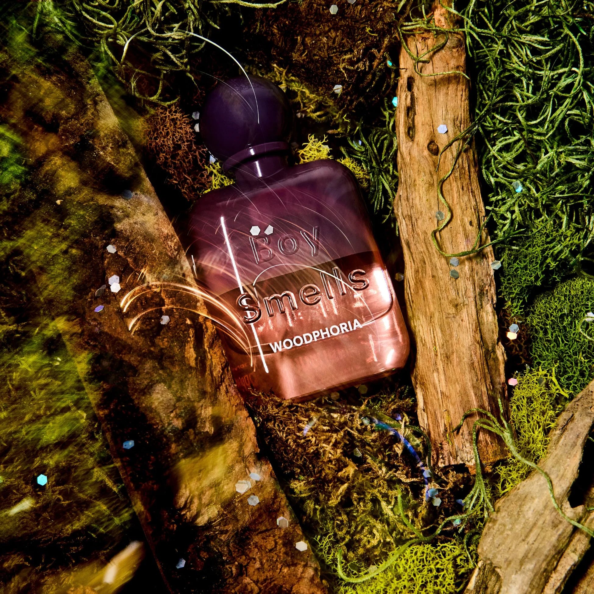 Boy Smells “Woodphoria” fragrance bottle nestled among moss, wood, and greenery with scattered light reflections.
