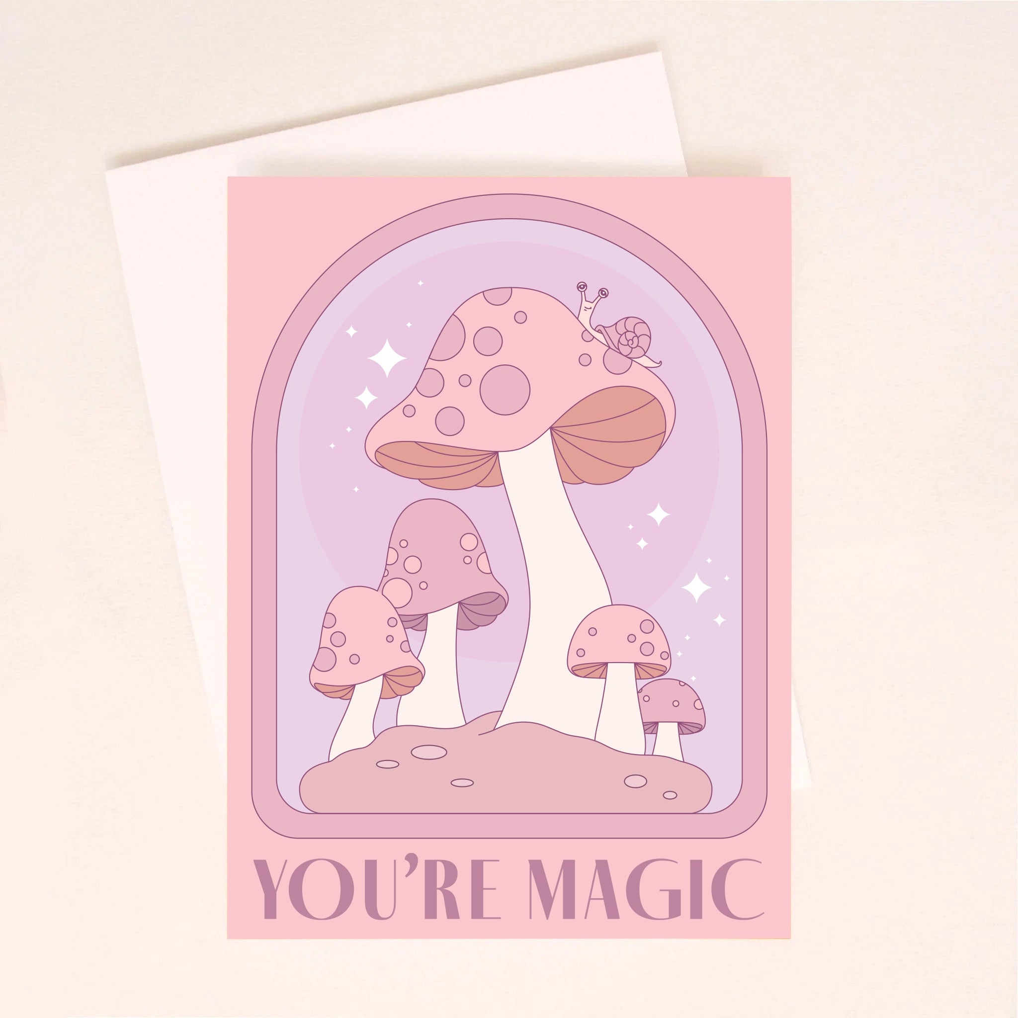 On a light pink background is a pink and purple card with an arch and mushroom design as well as text along the bottom that reads, &quot;You&#39;re Magic&quot;. A white envelope is included with purchase, also shown here.