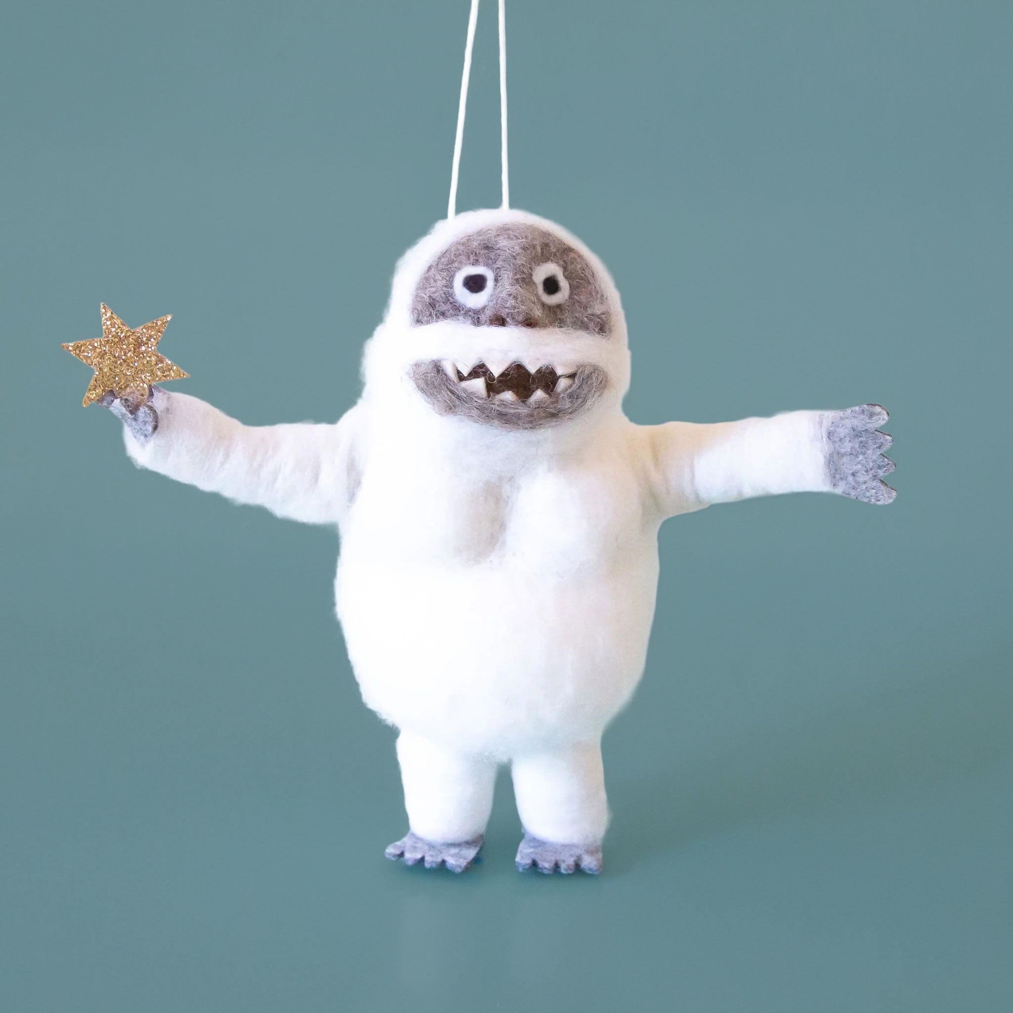 On a light blue background is a white and grey felt abominable snowman ornament that's holding a sparkly star