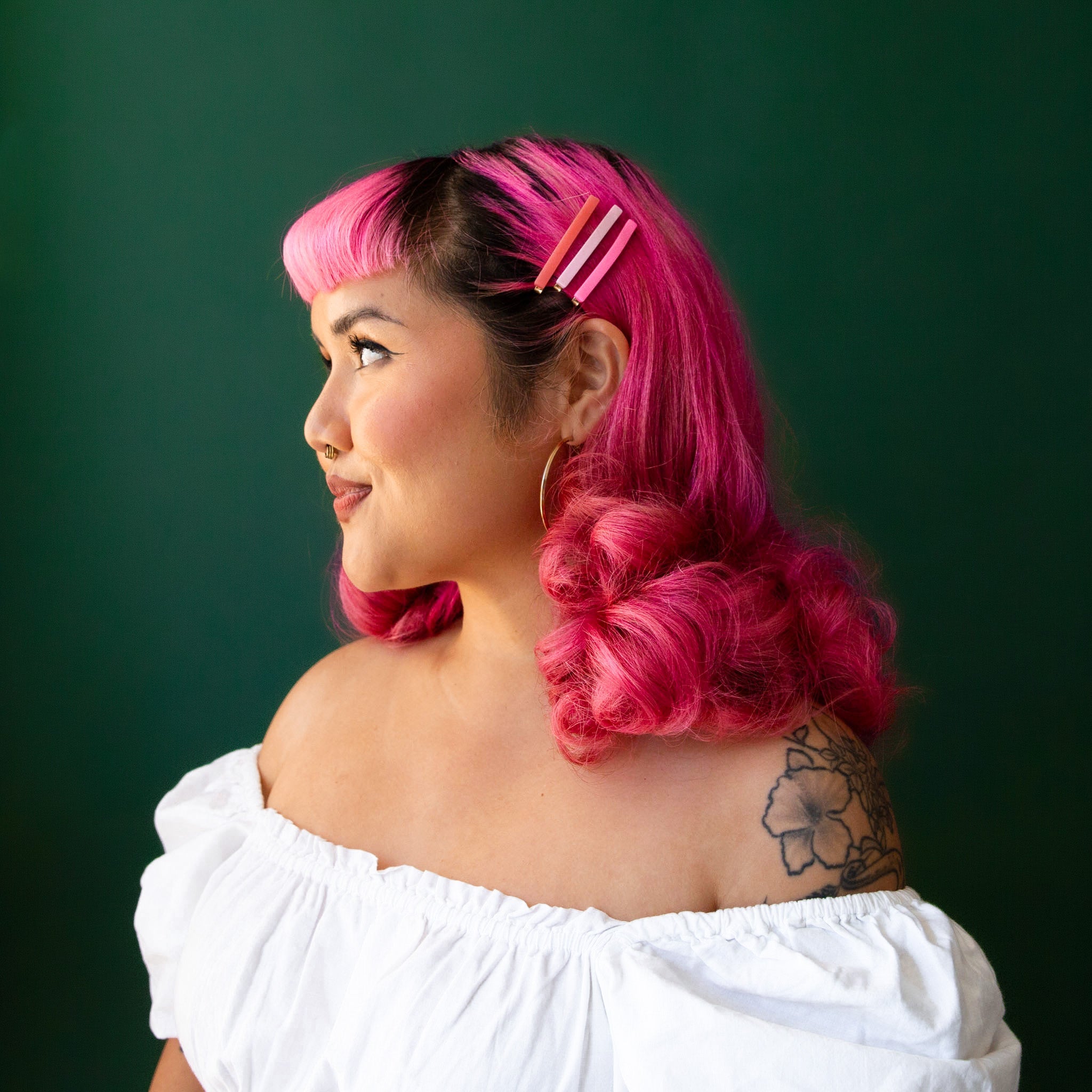 A model with pink hair and a white off the shoulder top wears three pink hair clips.  She is in front of an emerald green background.