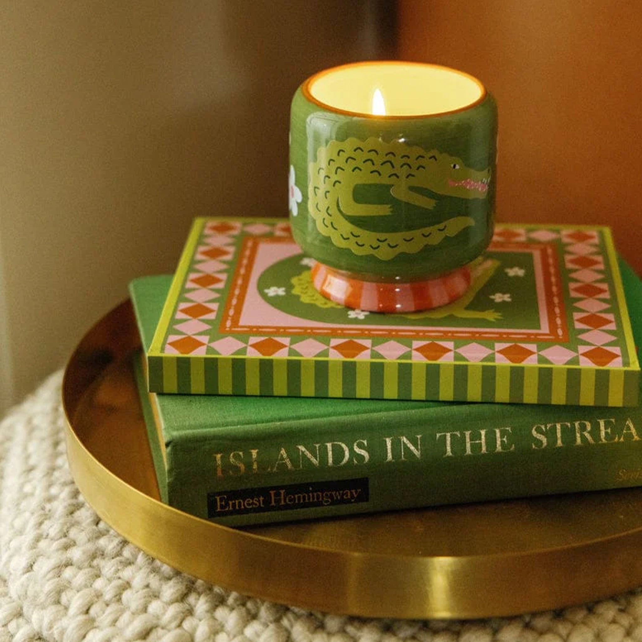 A green candle with a crocodile motif sits on top of two books in a gold tray.