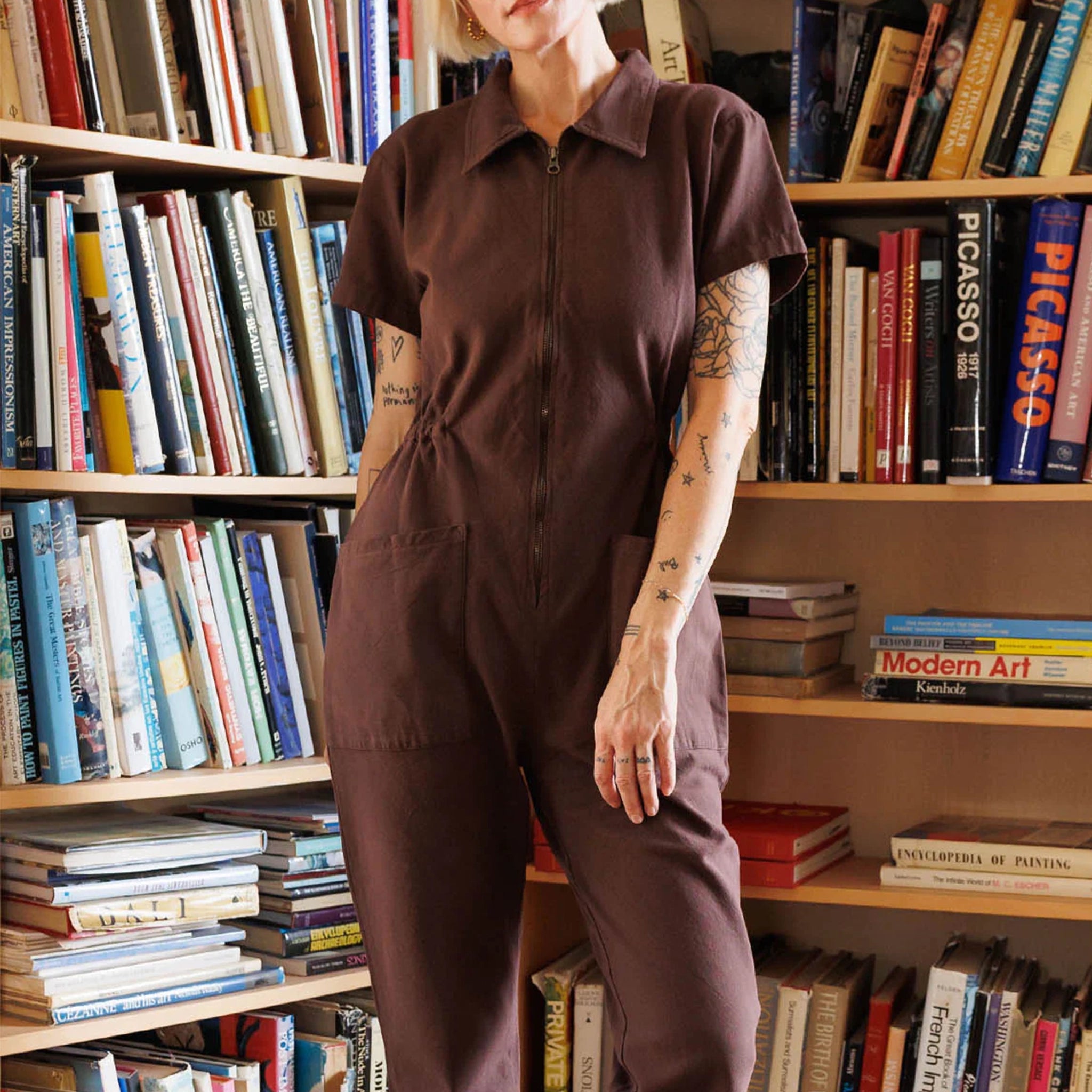 A model standing in front of a bookcase with a chocolate brown utility jumpsuit on with a zipper going up the front, a collar and a cinched waist.