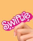 A pink and white air freshener out of its plastic packaging that reads, "Swiftie".