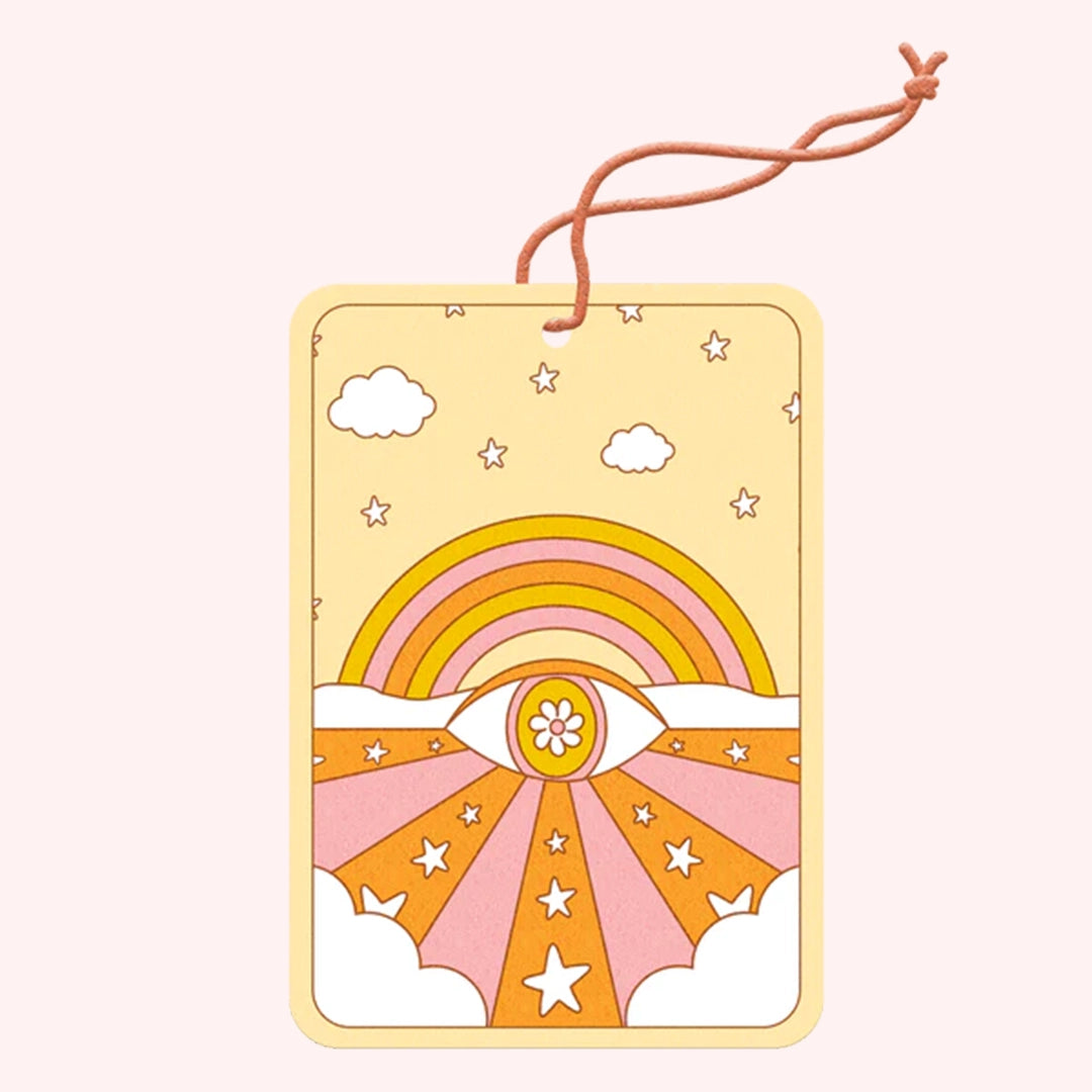 On a light pink background is a rectangle air freshener with an elastic loop for hanging and an orange, yellow and pink rainbow and all seeing eye illustration. 