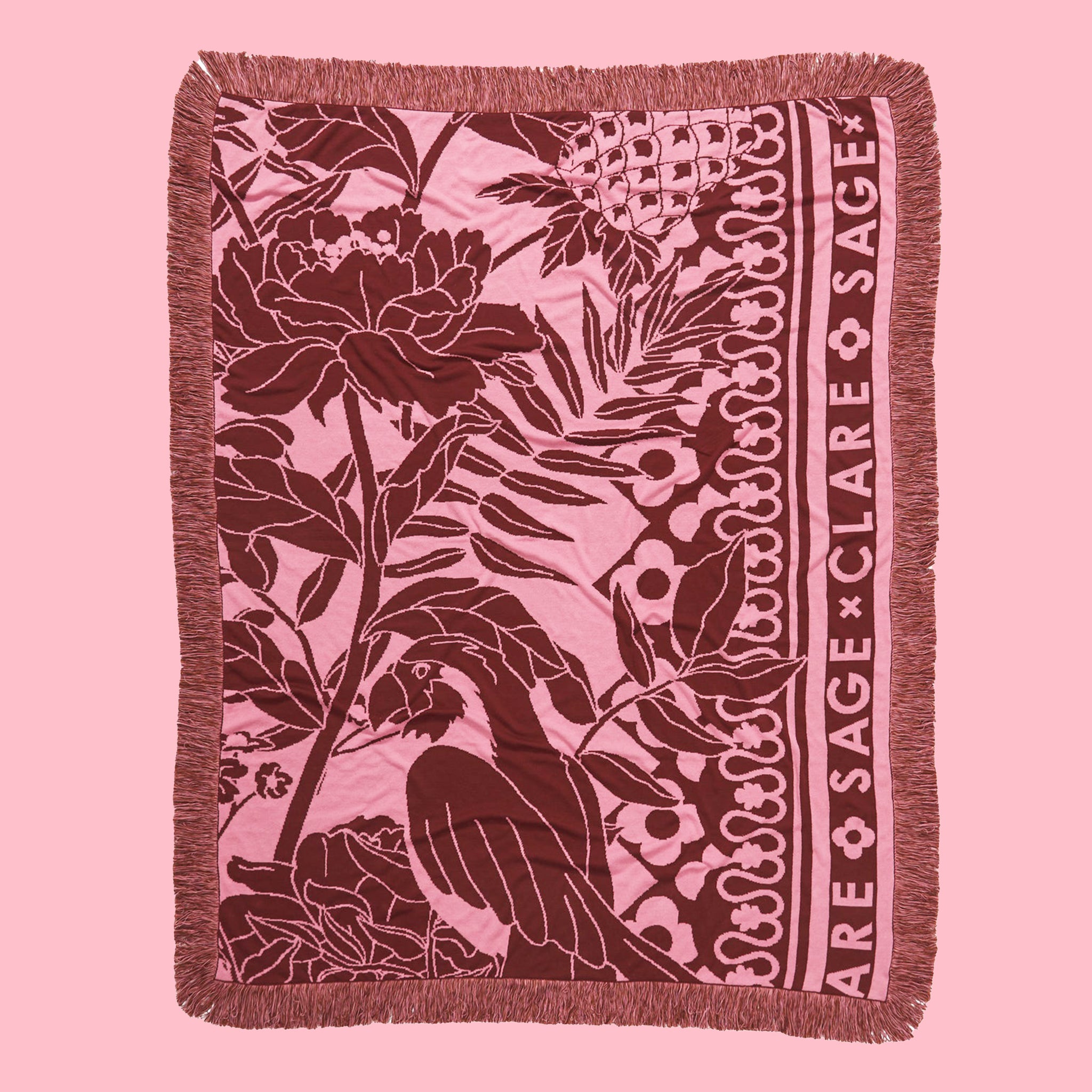 On a pink background is a pink and rust tropical print with &quot;sage x clare&quot; down the side of one edge along with fringe edges on all sides. 