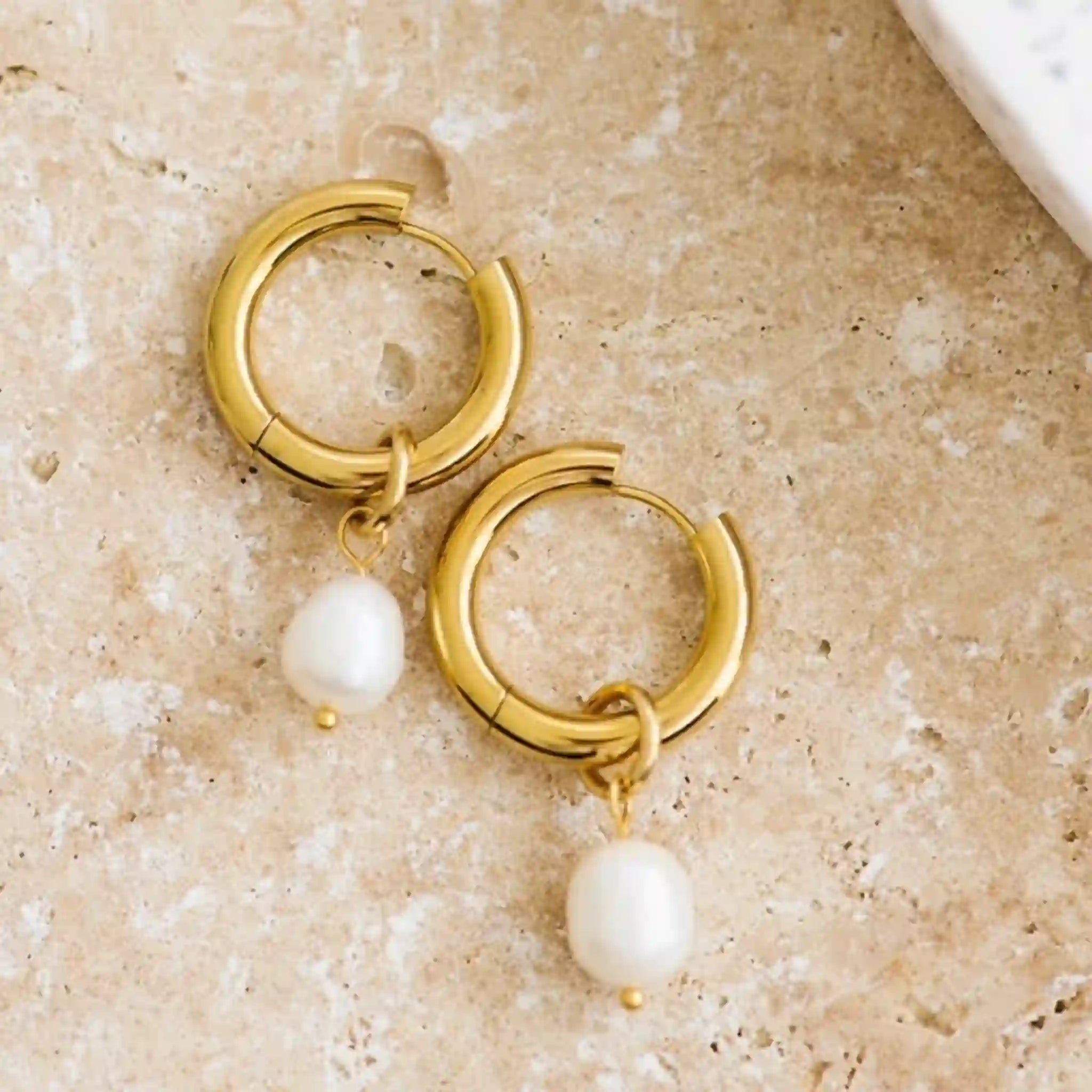 On a tan background is a pair of gold hoop earrings with a freshwater pearl dangling on each. 