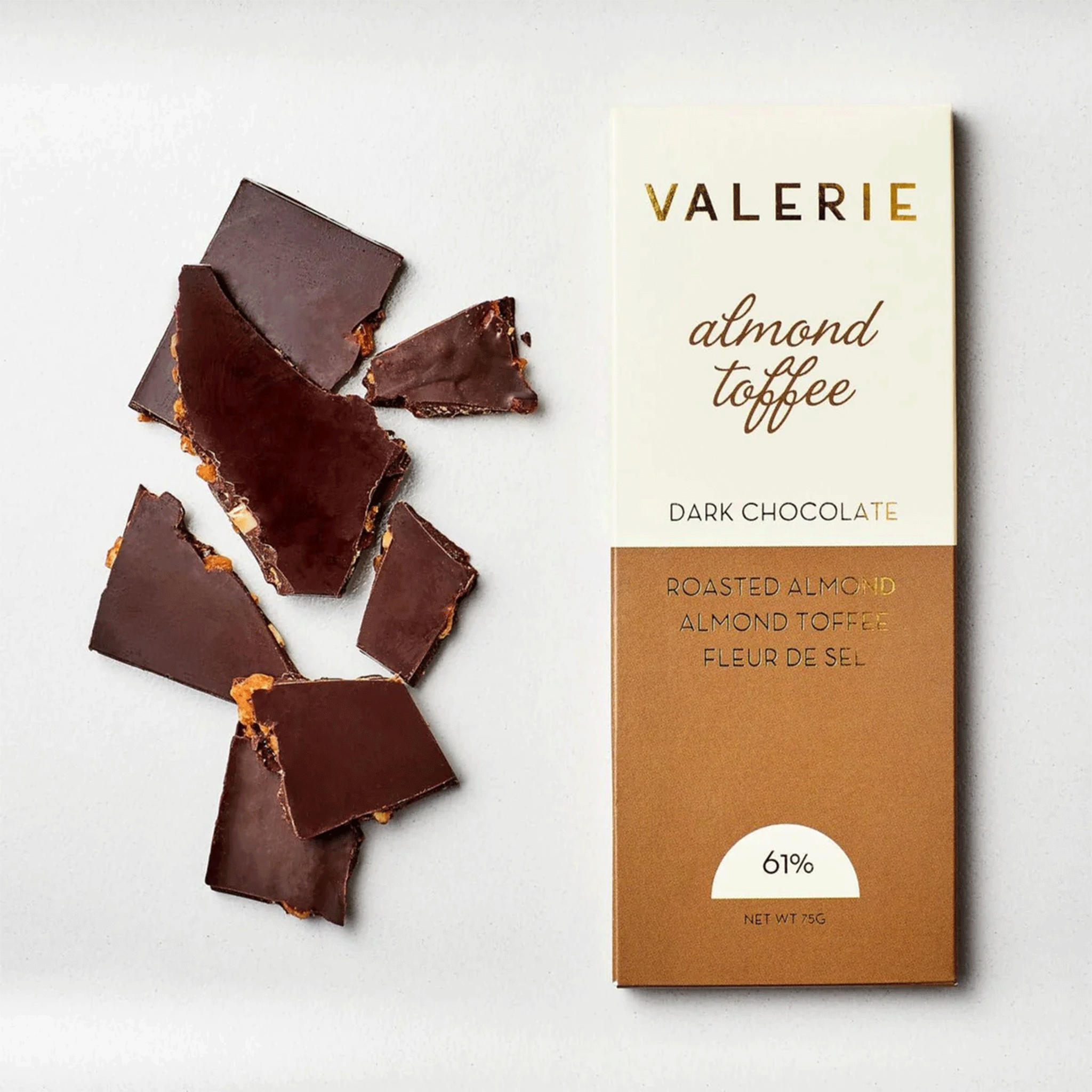 On a white background is a chocolate bar with tan and ivory packaging along with gold foil text that reads, &quot;Valerie almond toffee Dark Chocolate Roasted Almond Almond Toffee Fleur De Sel&quot;.