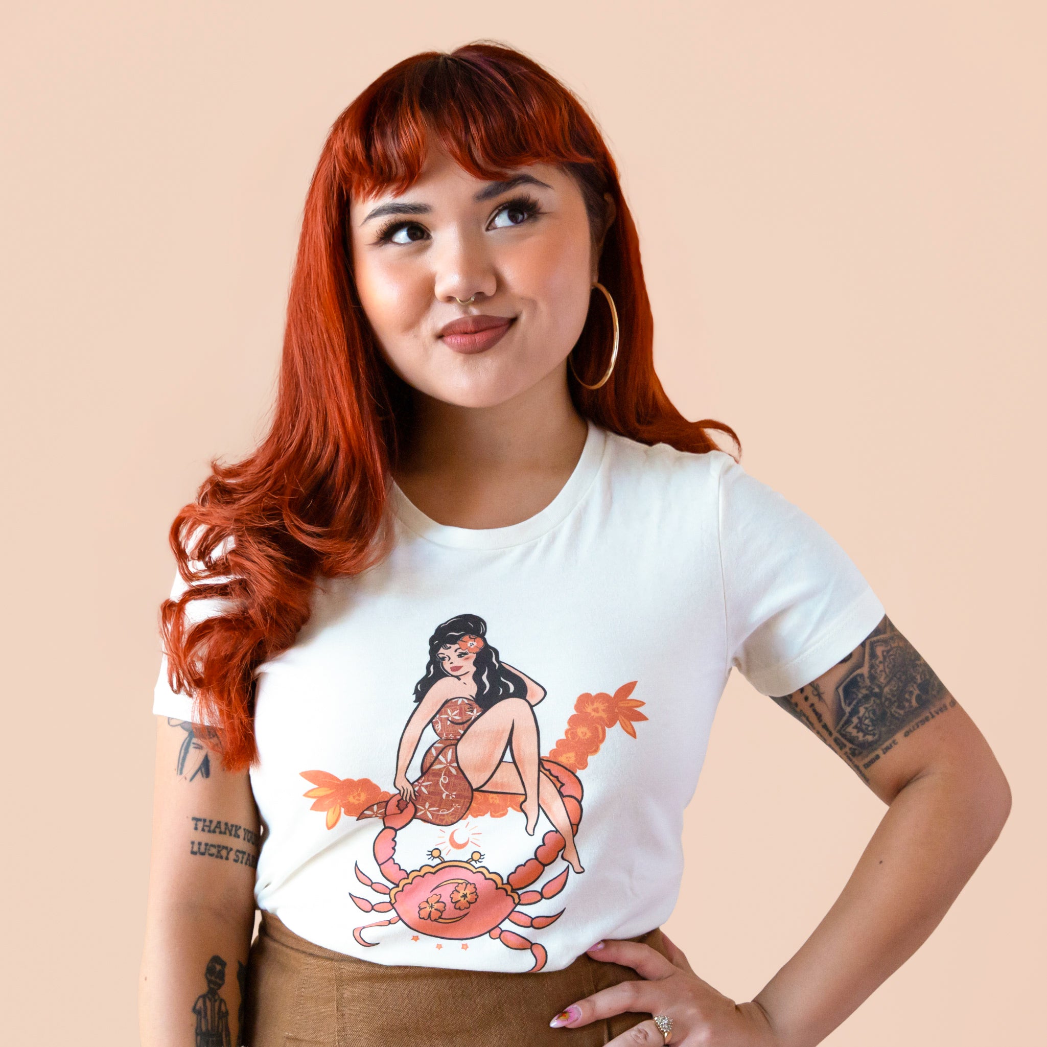 A model with red hair wearing an ivory tshirt with red piping details on the sleeves and neckline and a graphic of a vintage inspired woman with a hawaiian flower in her hair above a crab. 