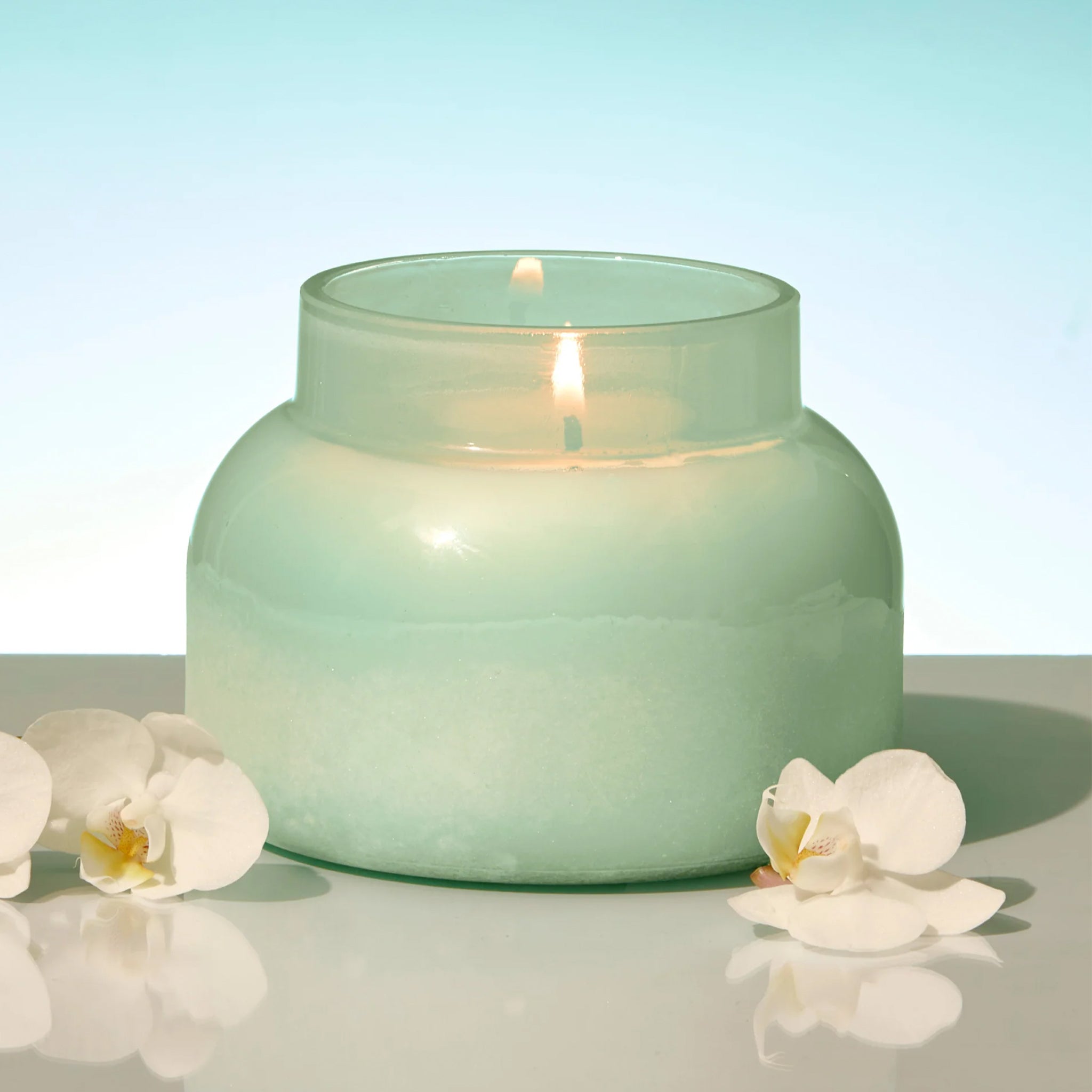 Green candle in a glass jar with white flowers on a reflective surface