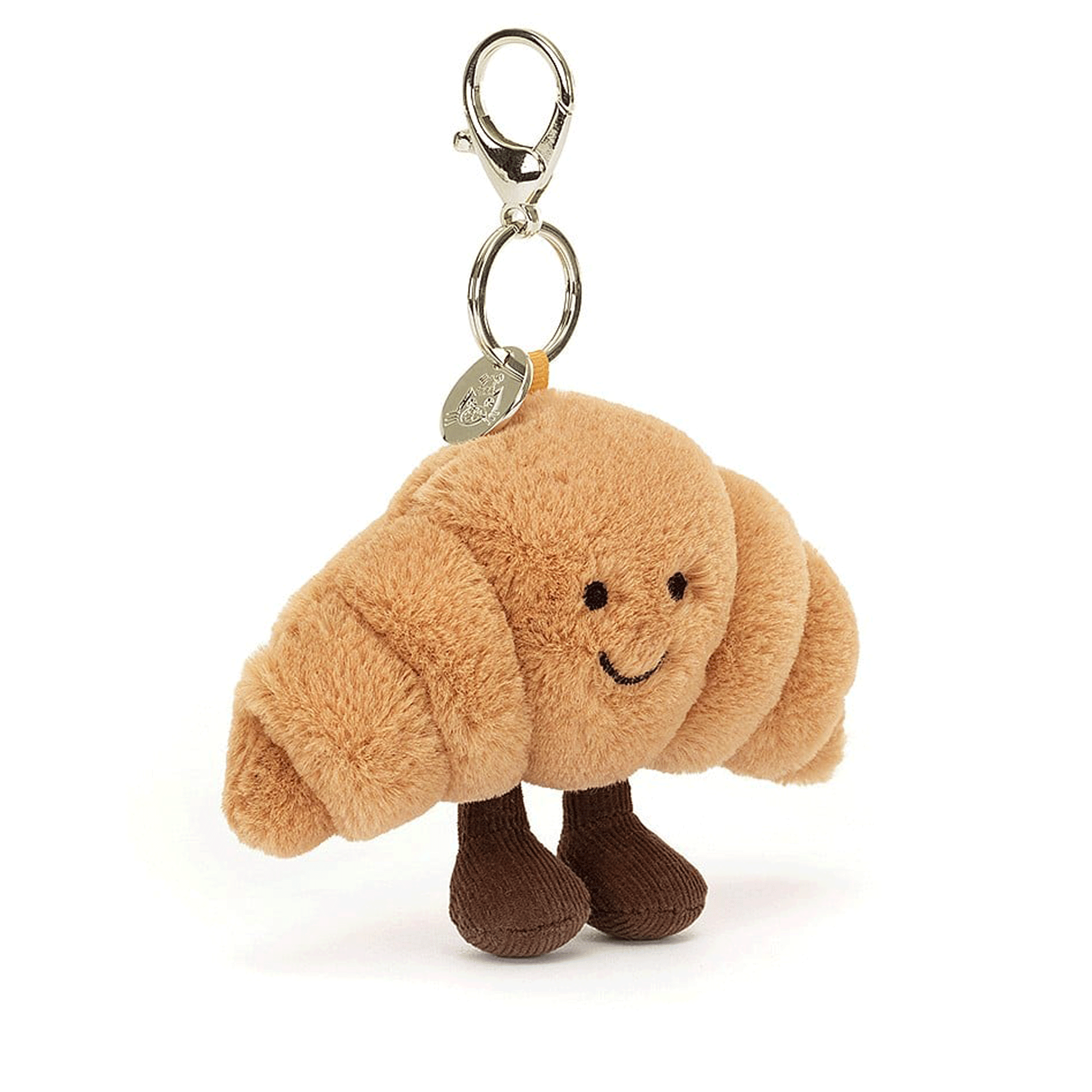 On a white background is a tan colored croissant stuffed toy with a silver clasp for attaching to a bag. 