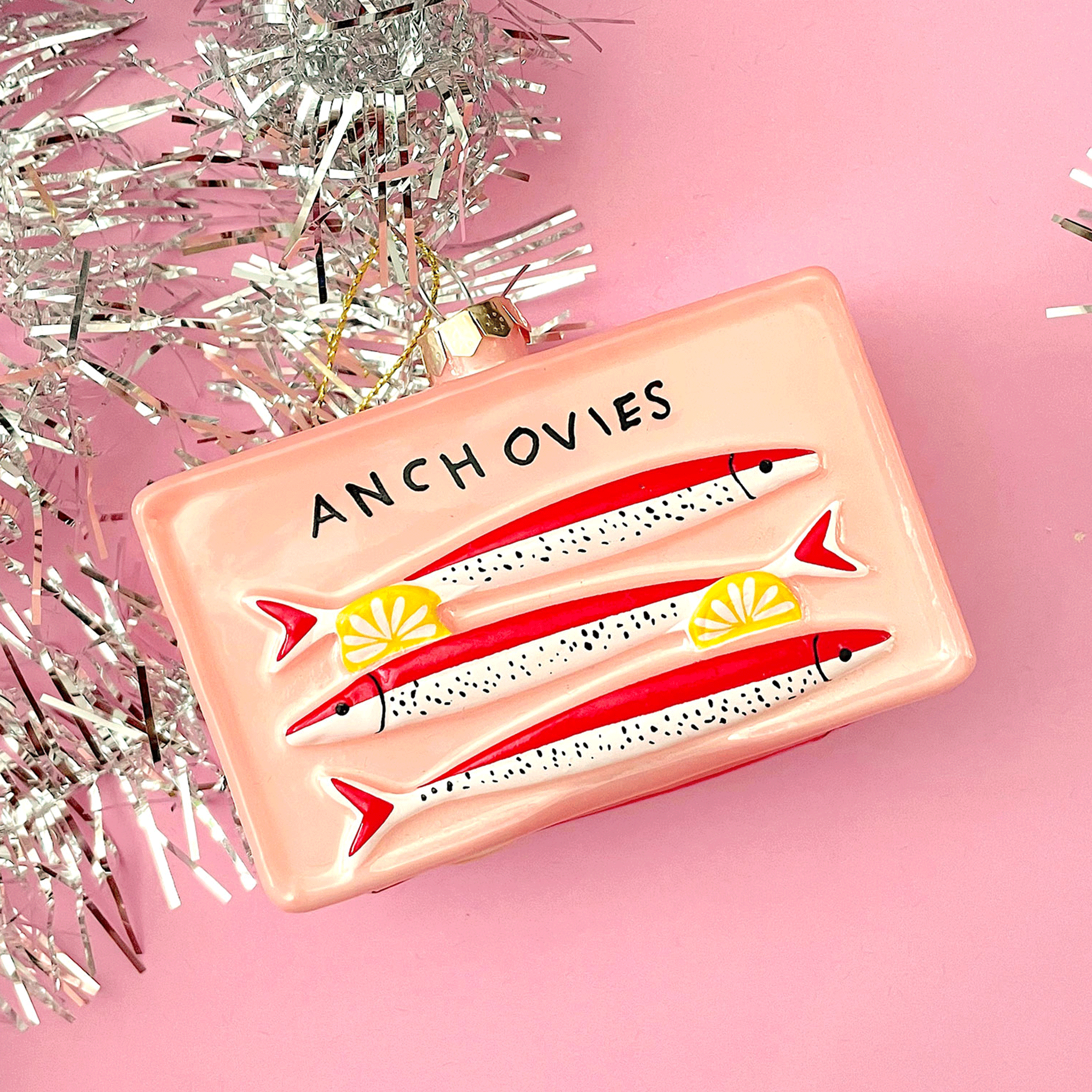 Christmas ornament in a pink rectangle shape with three illustrated white sardines with red backs, and two lemon slices, above is text "Anchovies"