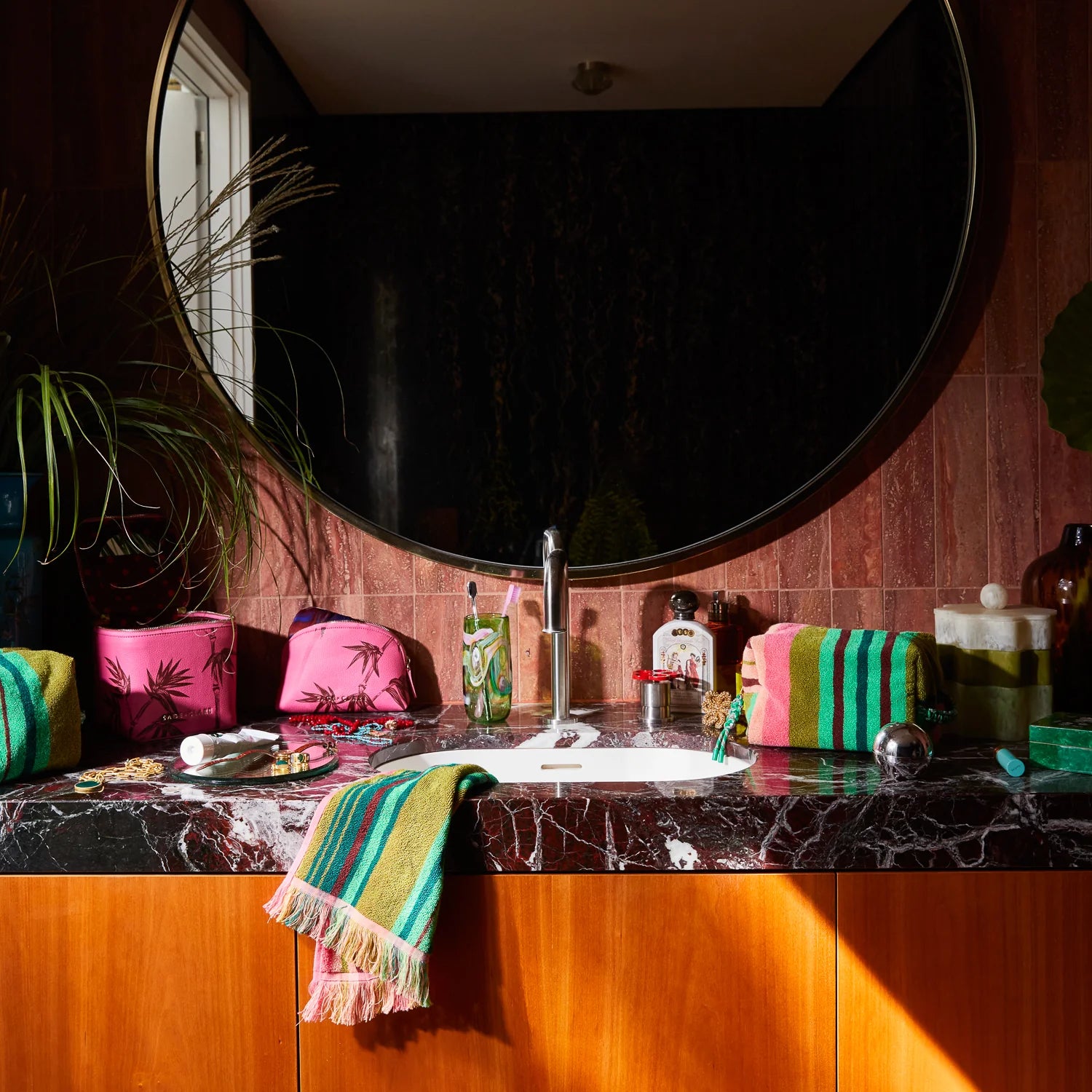 Bathroom vanity with a striped hand towel draped over the cabinet, surrounded by pink cosmetic pouches and assorted toiletries beneath a round mirror.