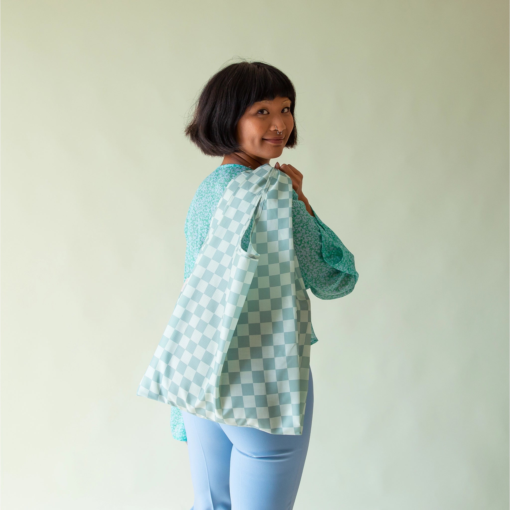 On a light blue background is a model holding a turquoise and light blue checkered print nylon bag.  