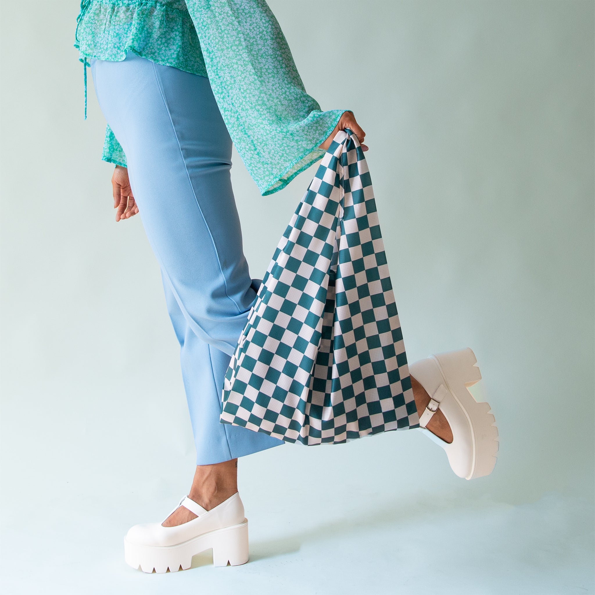 On a light blue background is a model holding a dark turquoise and light blue checkered print reusable nylon bag.