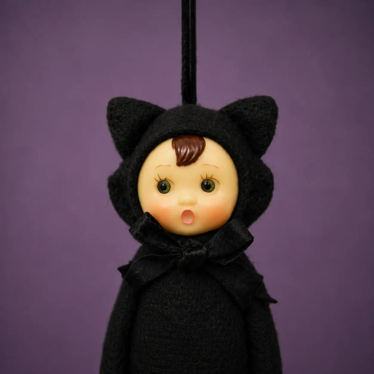 A black cat-cloaked doll with a round beige face, brown hair, and green eyes, hanging against a solid purple background.