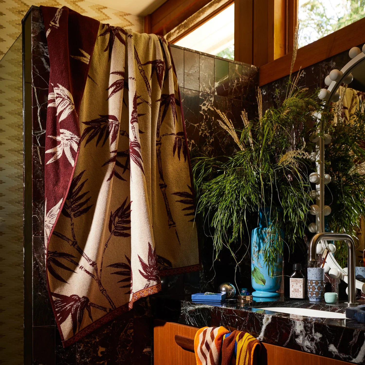 Neutral-toned towel with a leaf pattern draped over a tiled bathroom partition beside a sink with plants and decor.