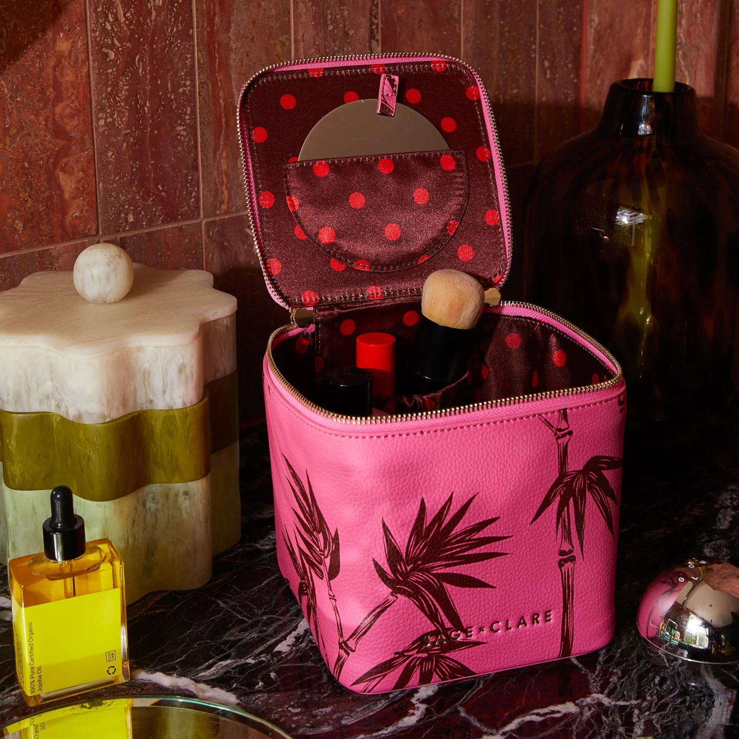 Open pink cosmetic case with palm print showing polka dot interior and makeup items on a marble bathroom counter.