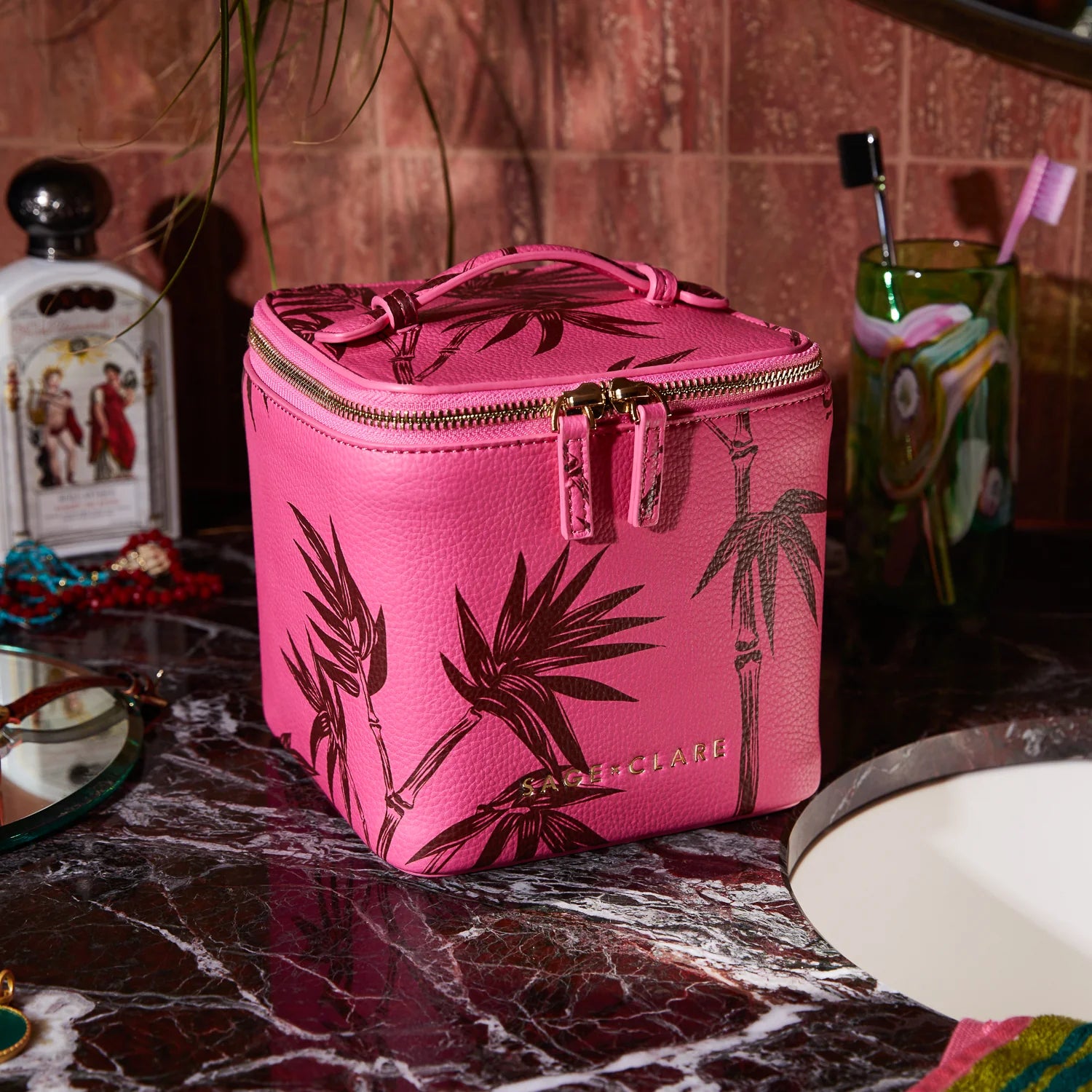 Pink cosmetic case with a palm print and top handle on a marble bathroom counter beside a sink and toiletries.
