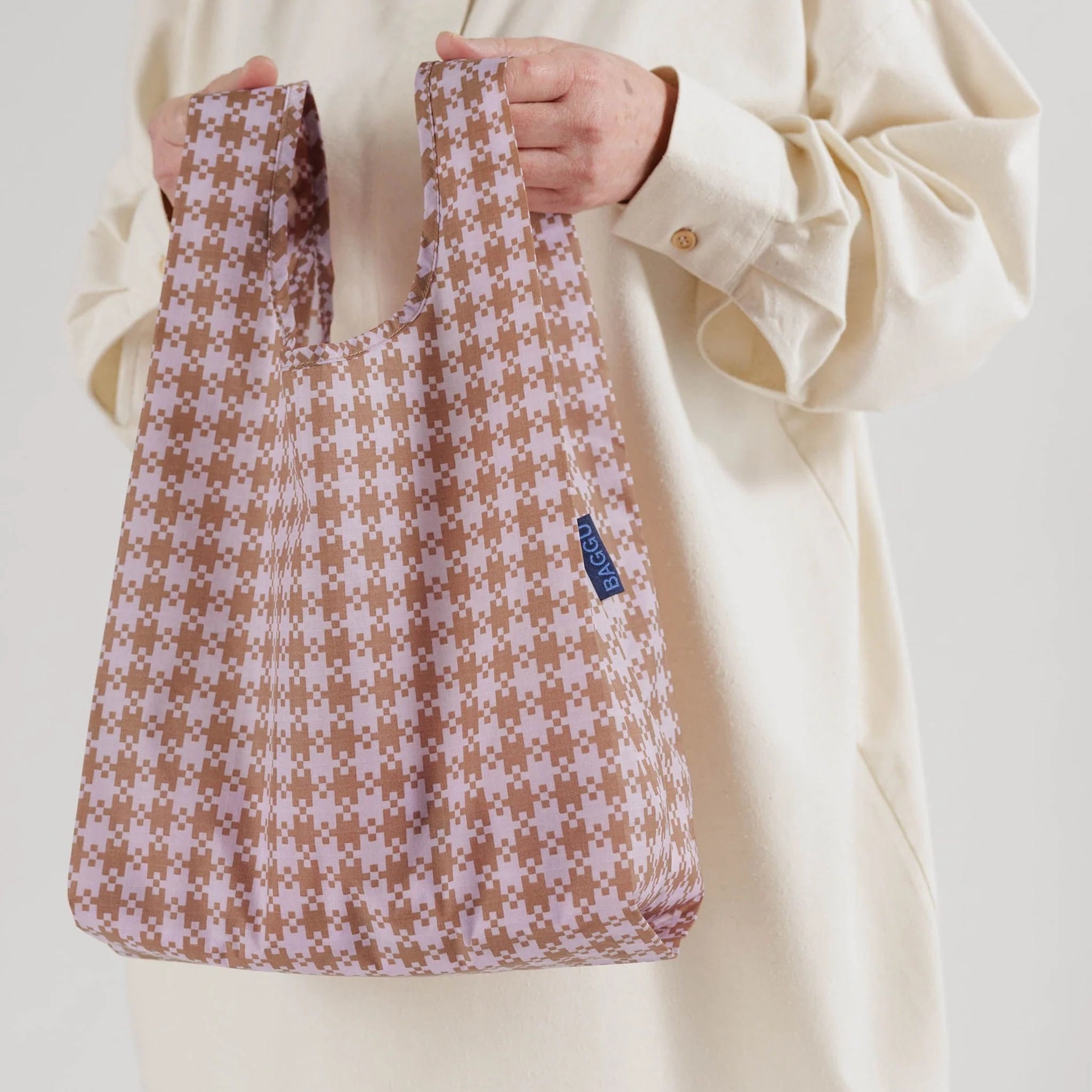 On a white background is a model holding a pink and tan gingham print tote bag made out of nylon. 
