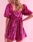On a pink background is a hot pink sequin romper with puff sleeves and a square neckline.