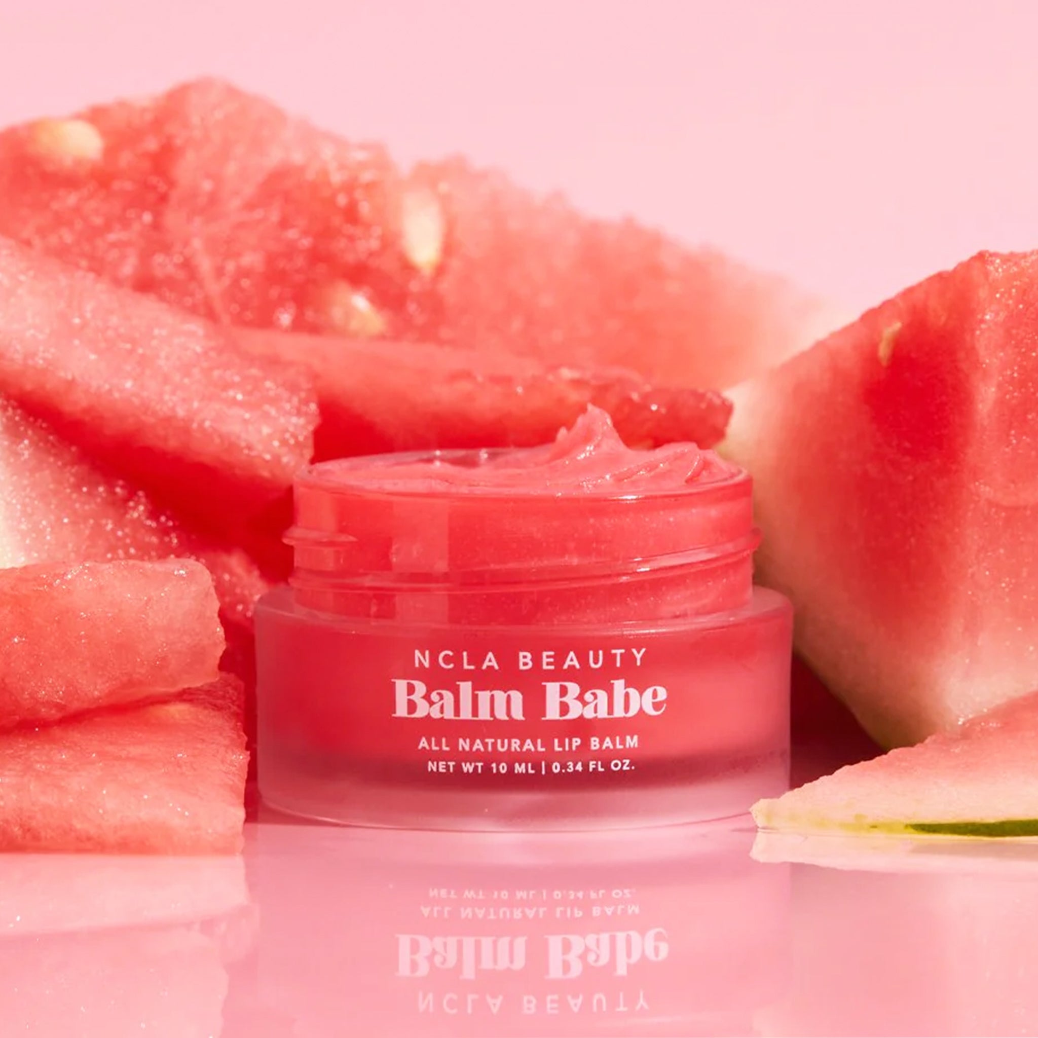 On a pink background is a small jar of pink lip balm surrounded by watermelon and white text across the front that reads, &quot;NCLA Beauty Balm Babe All Natural Lip Balm&quot;. 