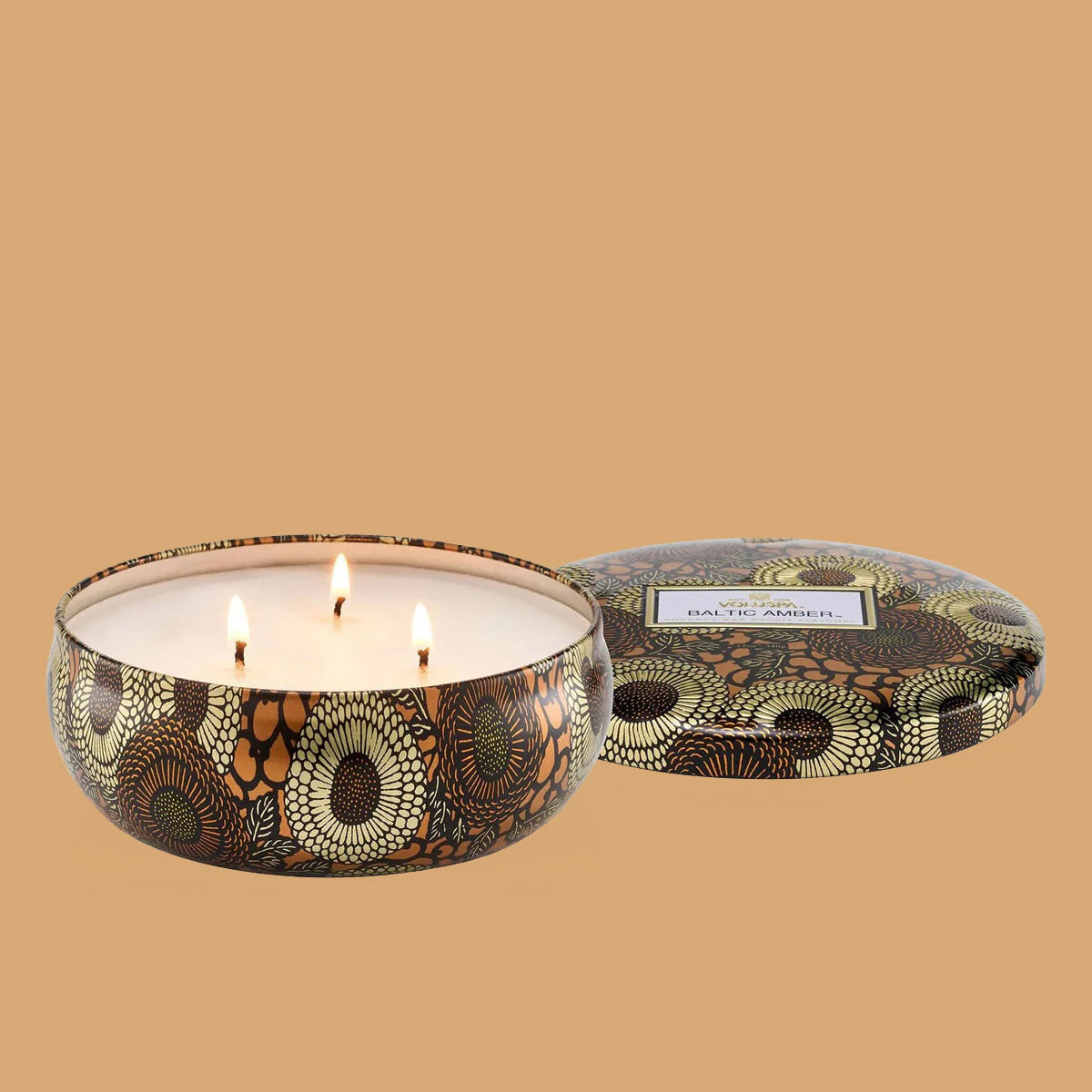 Round tin candle vessel with dark and light gold floral pattern and white wax candle with three wicks. Candle vessel lid has text, "Voluspa. Baltic Amber."