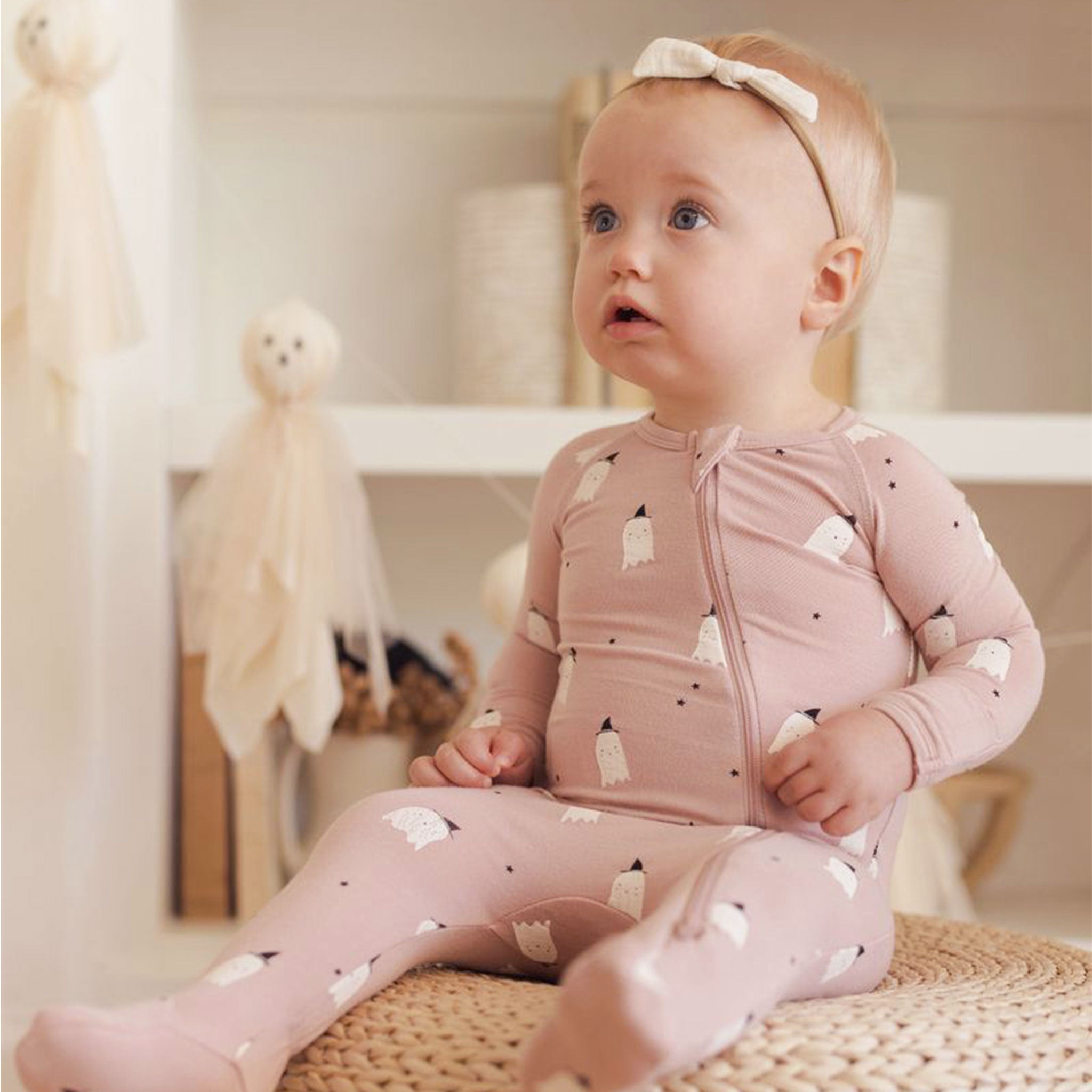 Baby wearing a pink footed onesie. The print is small white ghosts with black witch hats. The onesie has a zipper.