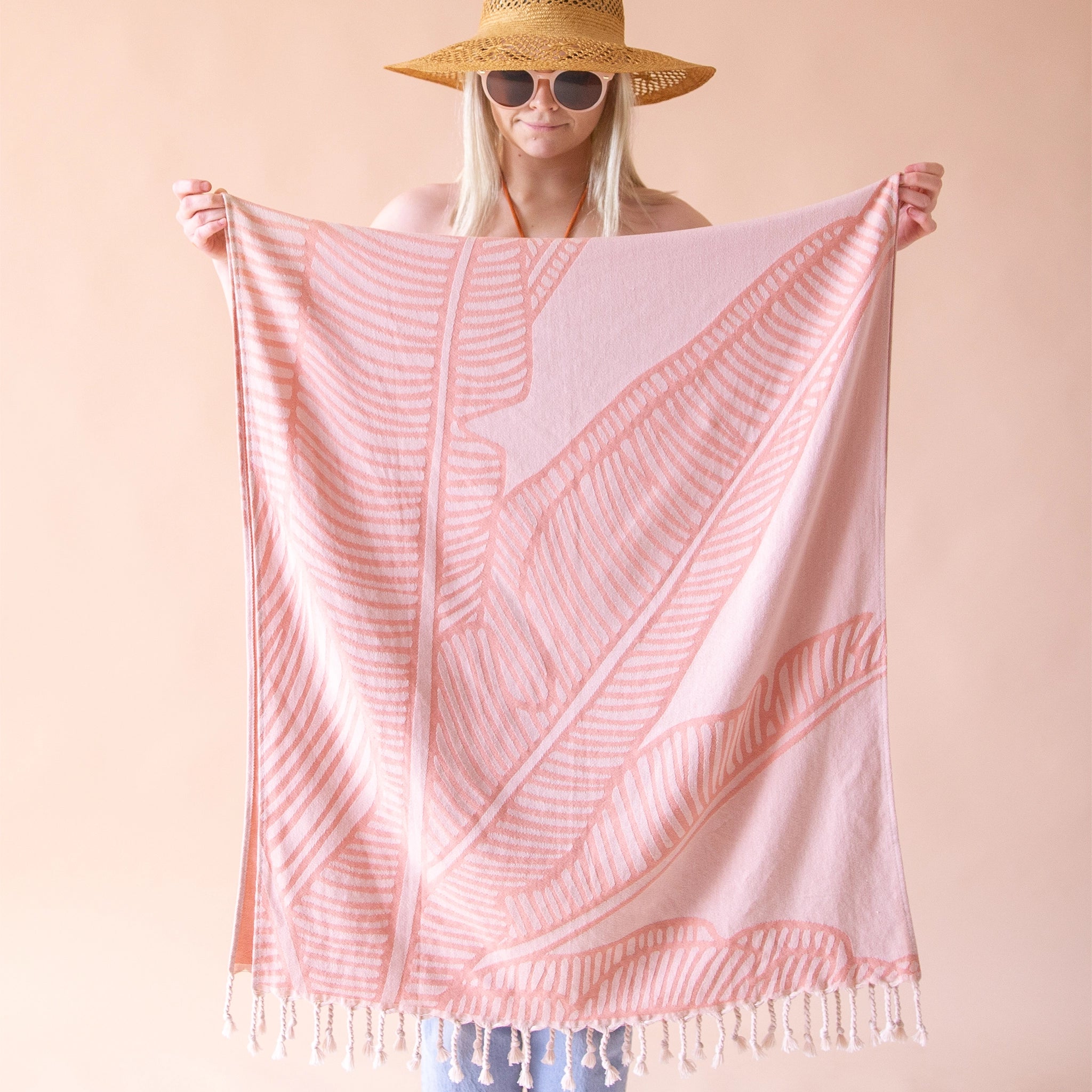 On a beige background is a model holding up the Bird of Paradise Beach Towel with the bird of paradise leaf print and string tassels on each end.