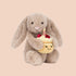 A beige bunny stuffed animal holding a birthday cake both with a smiling face. 