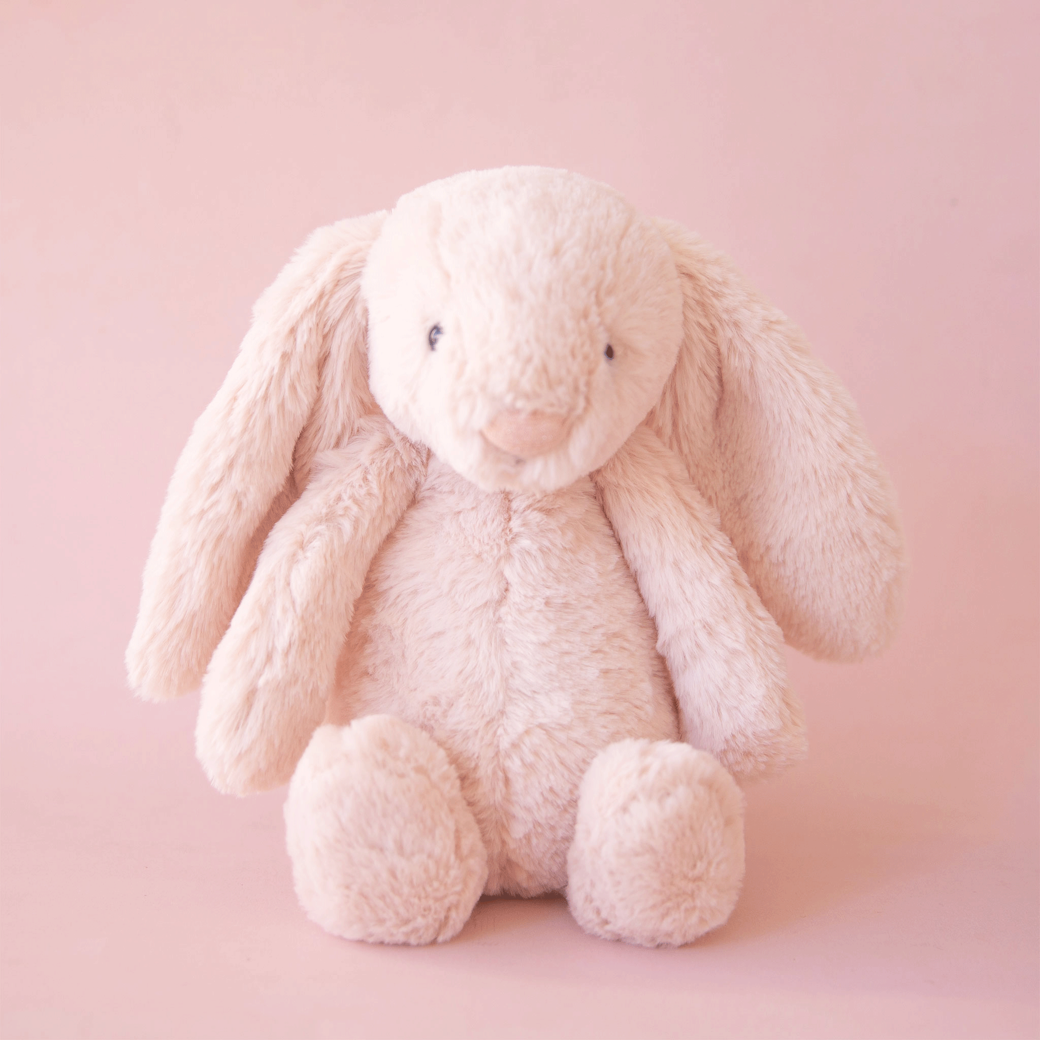 On a pink background is a cream colored stuffed animal bunny with long floppy ears and super soft faux fur.