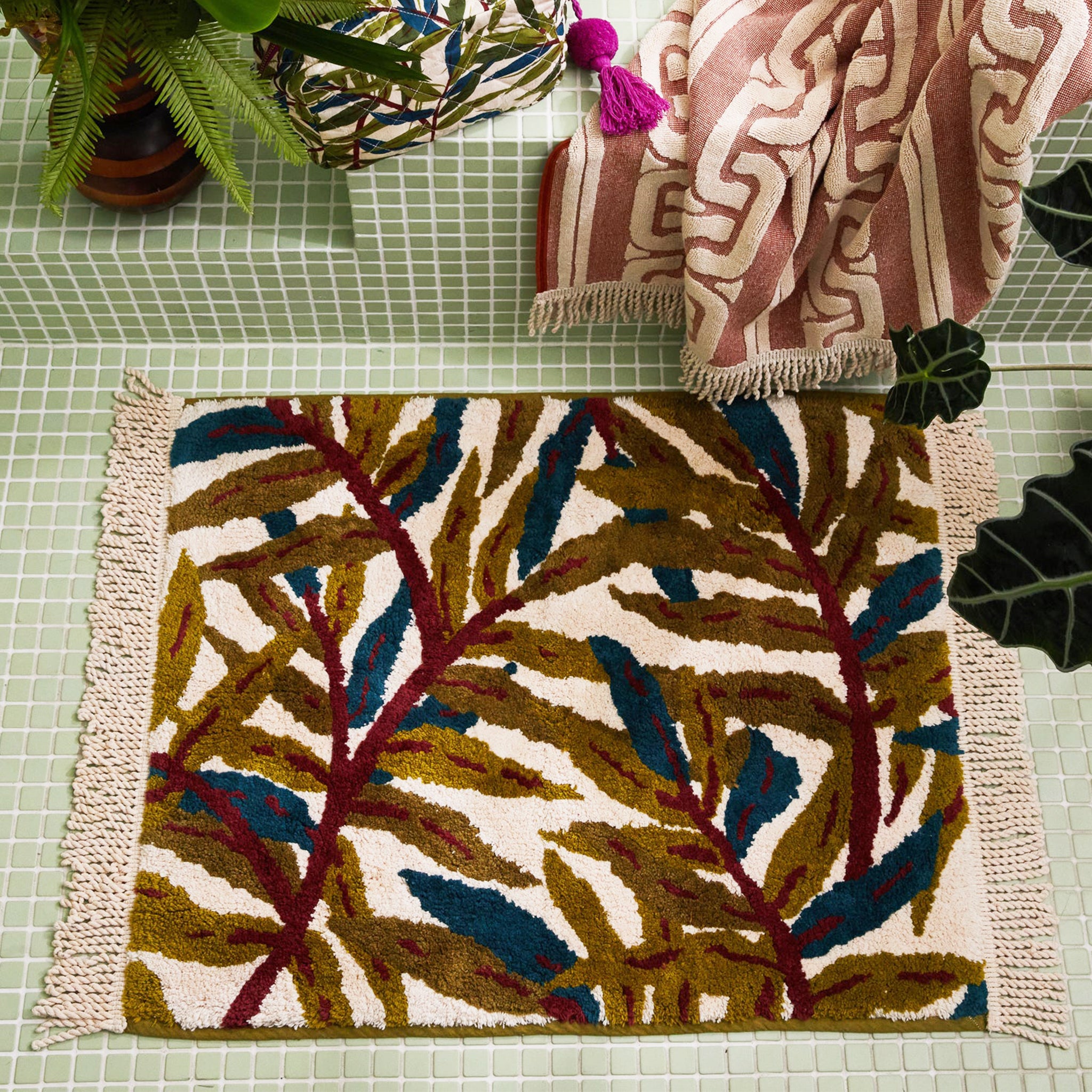A green, teal and ivory tropical foliage printed bath mat with tassel details on each end.