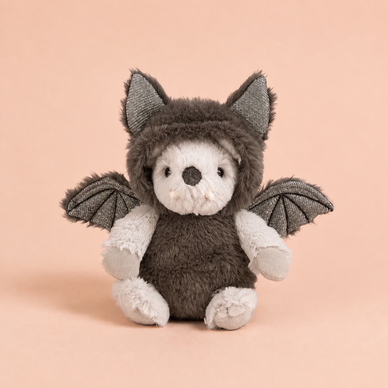 A plush toy styled as a bat, featuring a dark gray fuzzy body, white muzzle and paws, and gray wings, sitting against a solid peach-pink background.