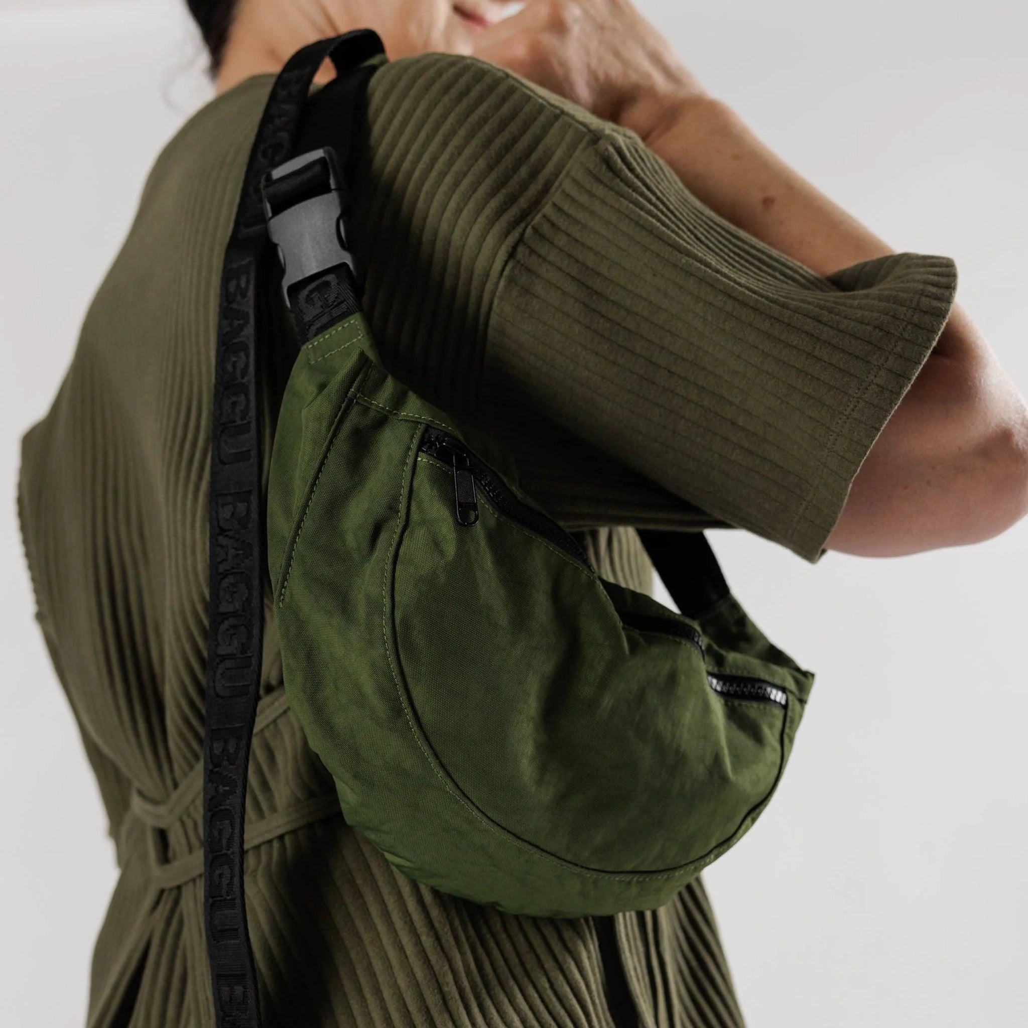 On a white background is a model wearing the dark green crescent shaped fanny pack with a black adjustable strap over their shoulder. 