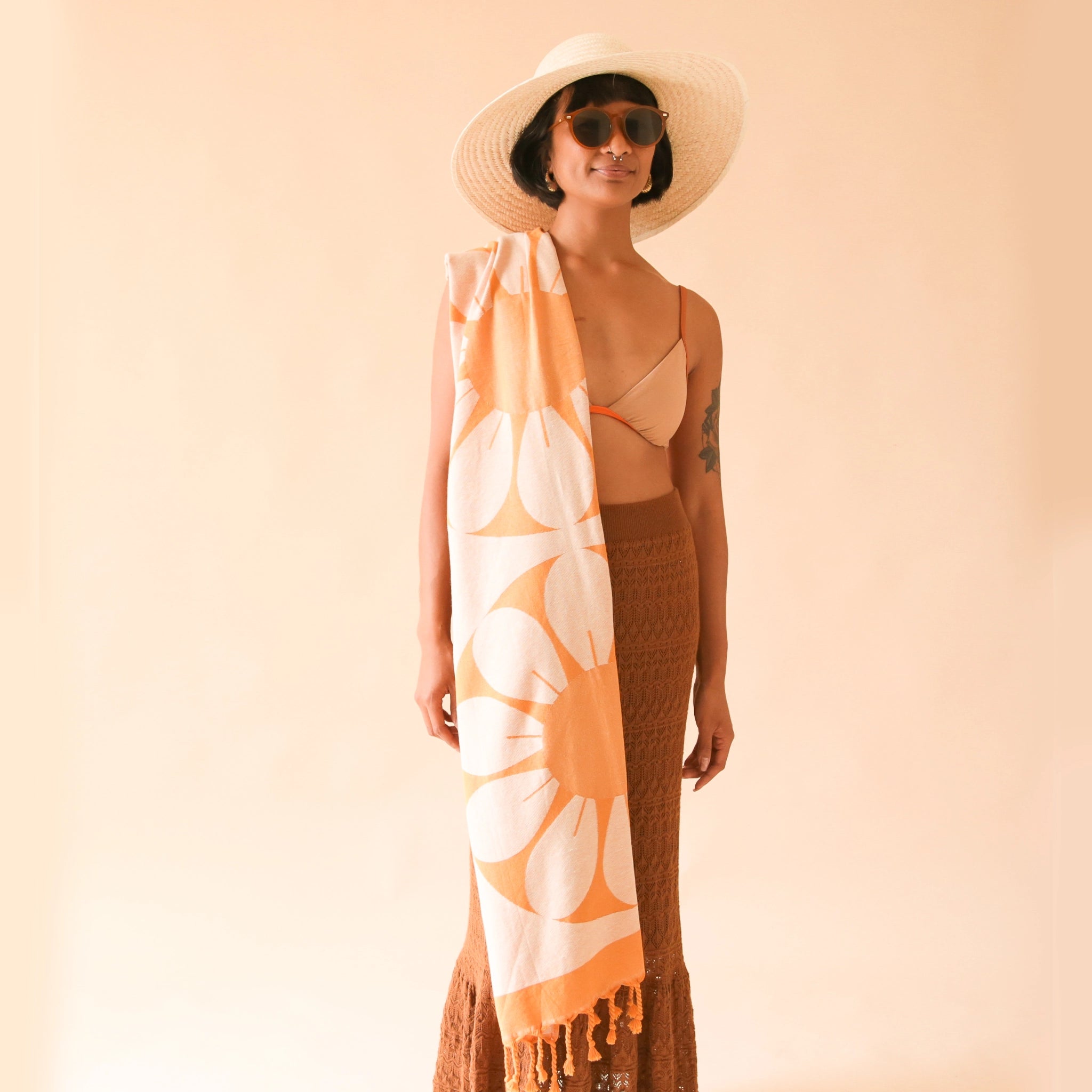 A model with the orange Retro Flower Beach Towel draped across their shoulder. The towel has two large flower designs and orange tassels at both ends. 