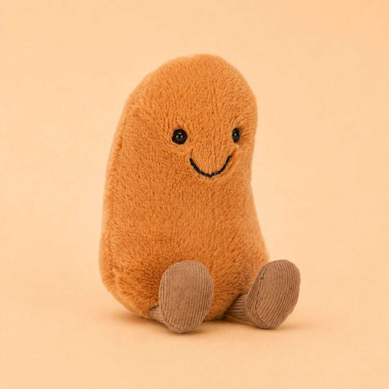 A plush bean-shaped toy in a warm tan-brown color with a simple black embroidered smiley face and gray-brown corduroy feet, sitting against a solid peach-beige background.