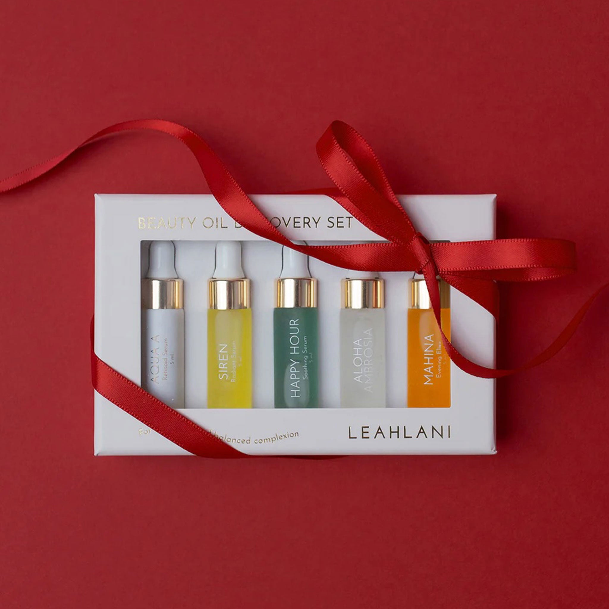 A white ‘Beauty Oil Discovery Set’ box from LEAHLANI sits on a deep red background, wrapped with a shiny red ribbon tied in a bow. Through a clear window, five small glass dropper bottles are visible, each with a gold collar and different colored oils/labels (light, yellow, green, clear, and orange). The arrangement feels festive and gift-ready.