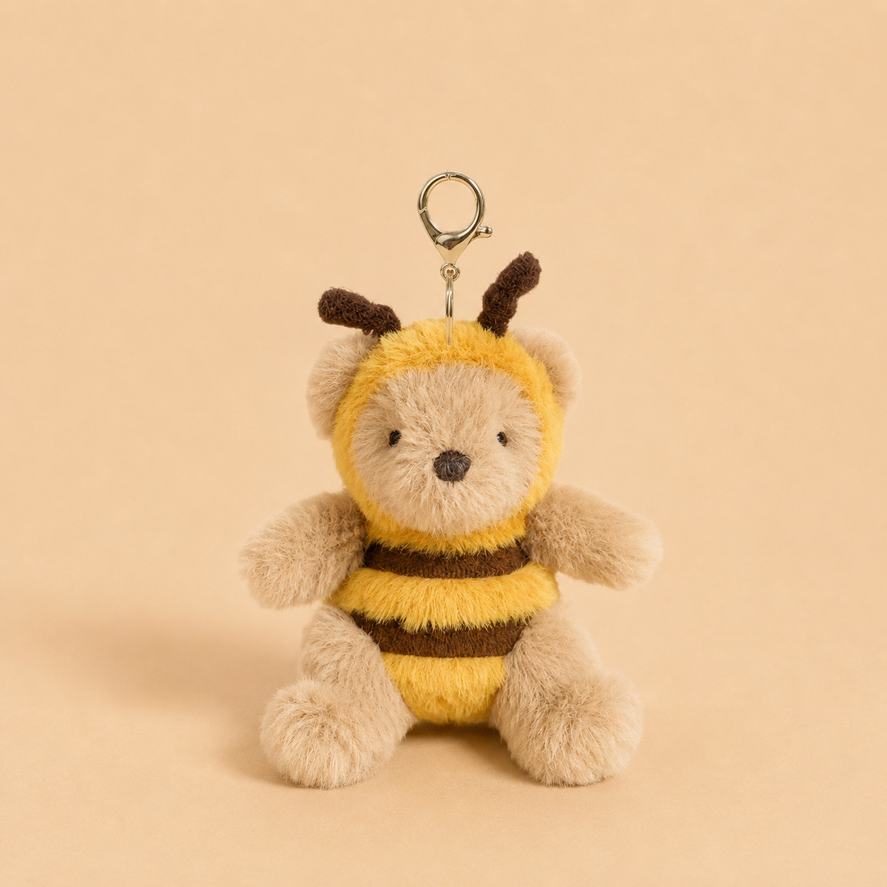 A plush teddy bear keychain dressed as a bee, featuring a soft beige body, yellow and brown striped fuzzy suit, and a silver metal clasp at the top, set against a solid warm beige background.