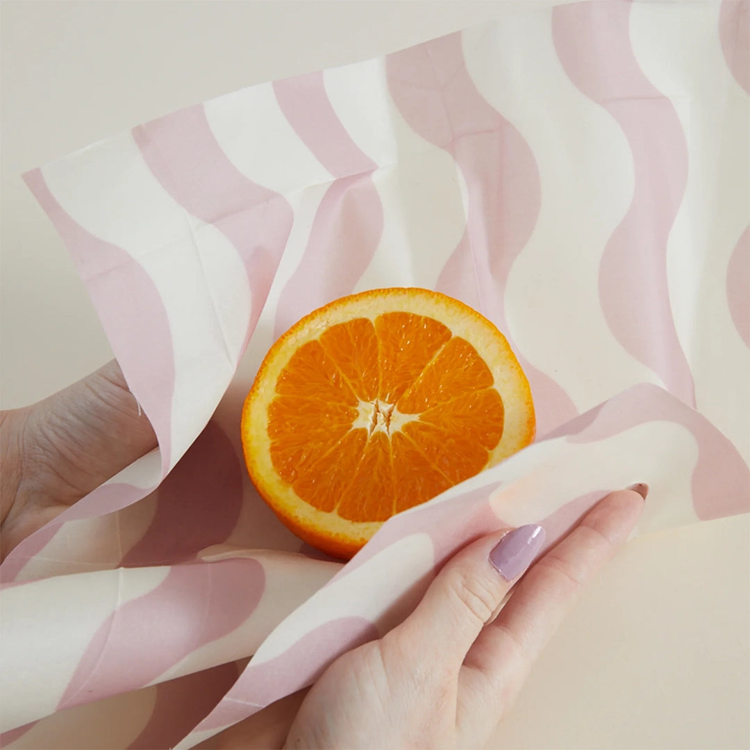 On a tan background is a piece of the beeswax wrap holding a cut orange. The wax wrap has a wavy pink and white design. 