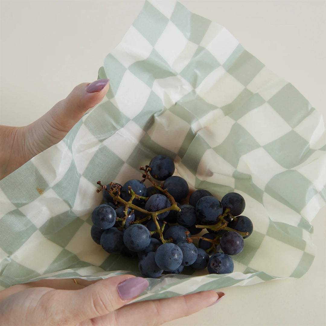 On a tan background is a piece of the beeswax wrap holding a vine of grapes. The wax wrap has a green and white checker pattern.  