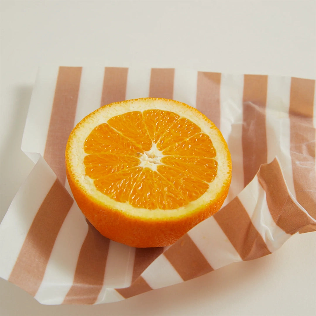 On a tan background is a piece of the beeswax wrap holding a cut orange. The wax wrap has an orange and white stripe design.