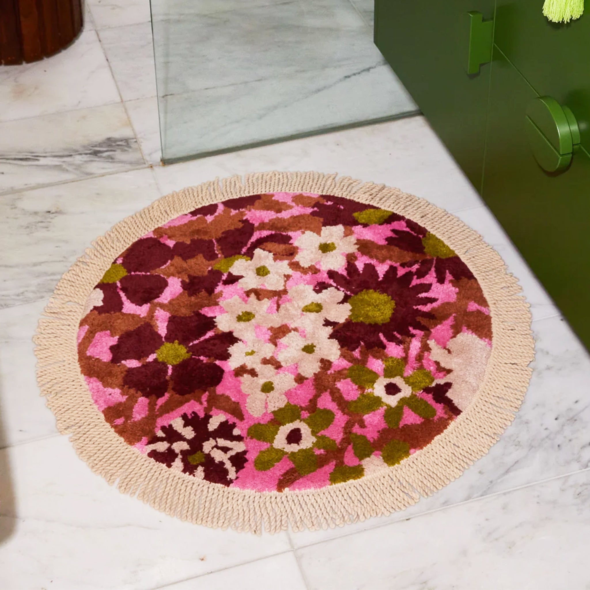 A pink, green and white floral print circular bath mat with a fringe detail around the edge. 
