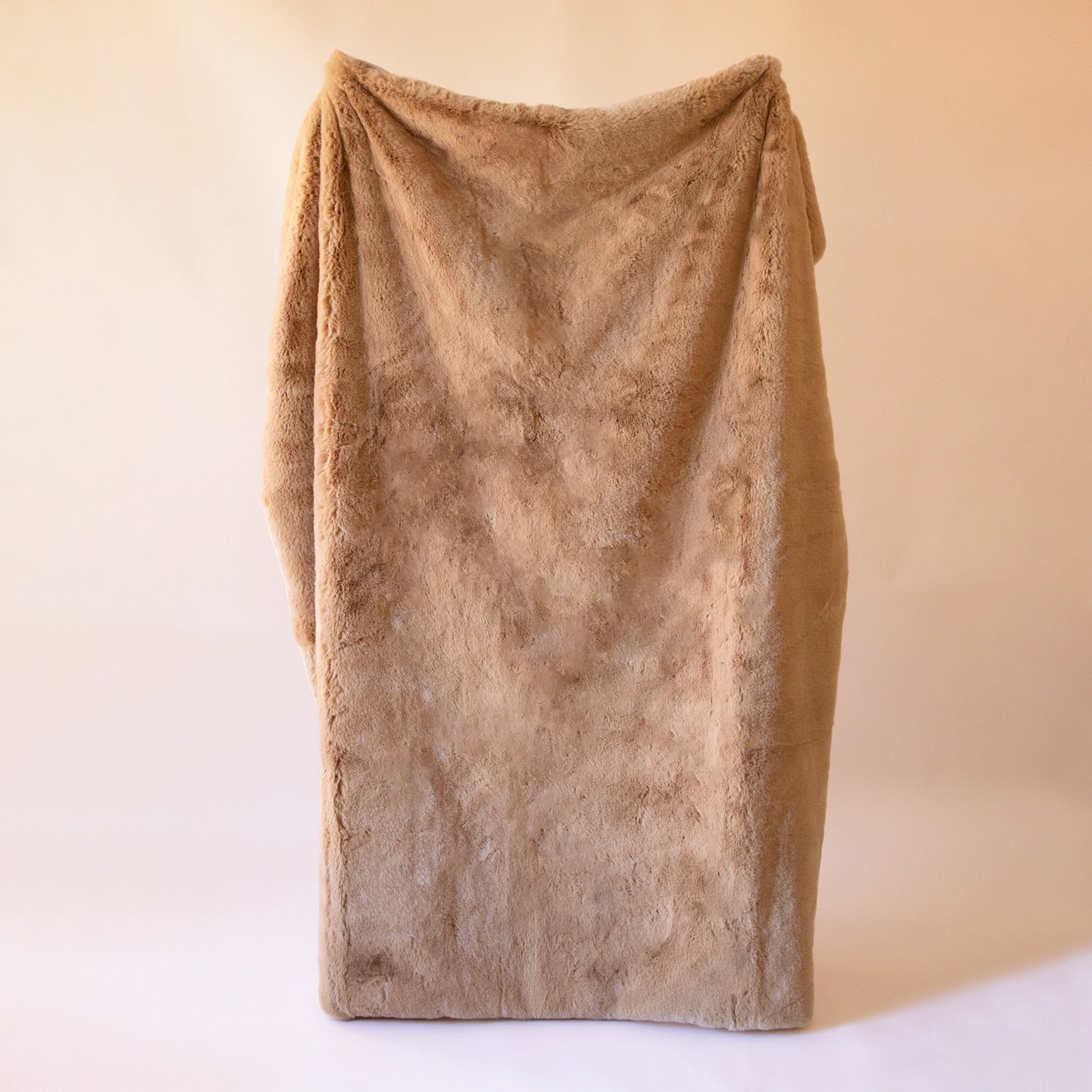 A large camel colored fur blanket being held by a person on a light tan background.