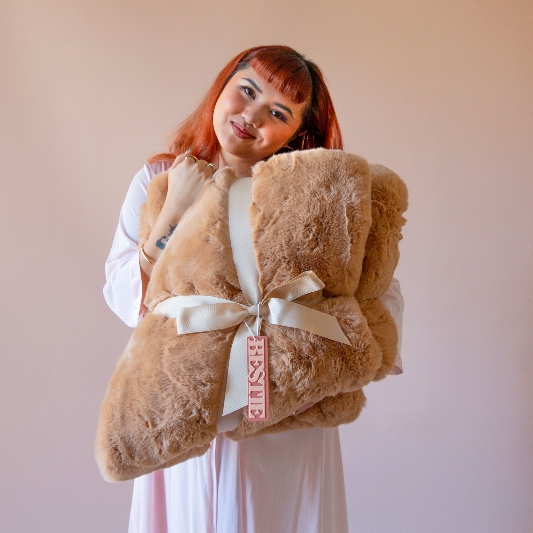 A red headed model holds a giant folded brown fur blanket with a satin bow.