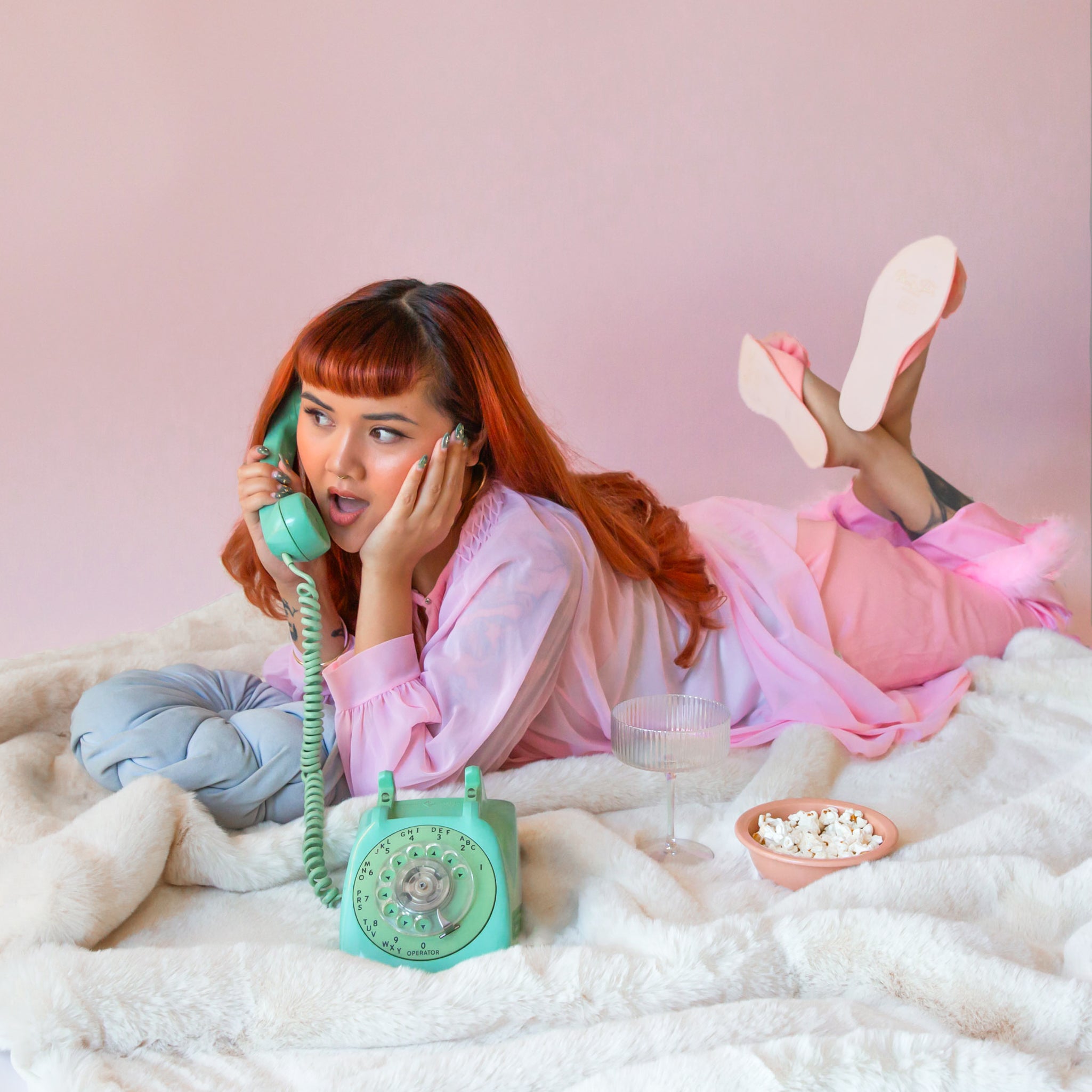 A girl on a mint telephone lays on top of a white fur blanket in pink pj's with a bowl of popcorn next to her.